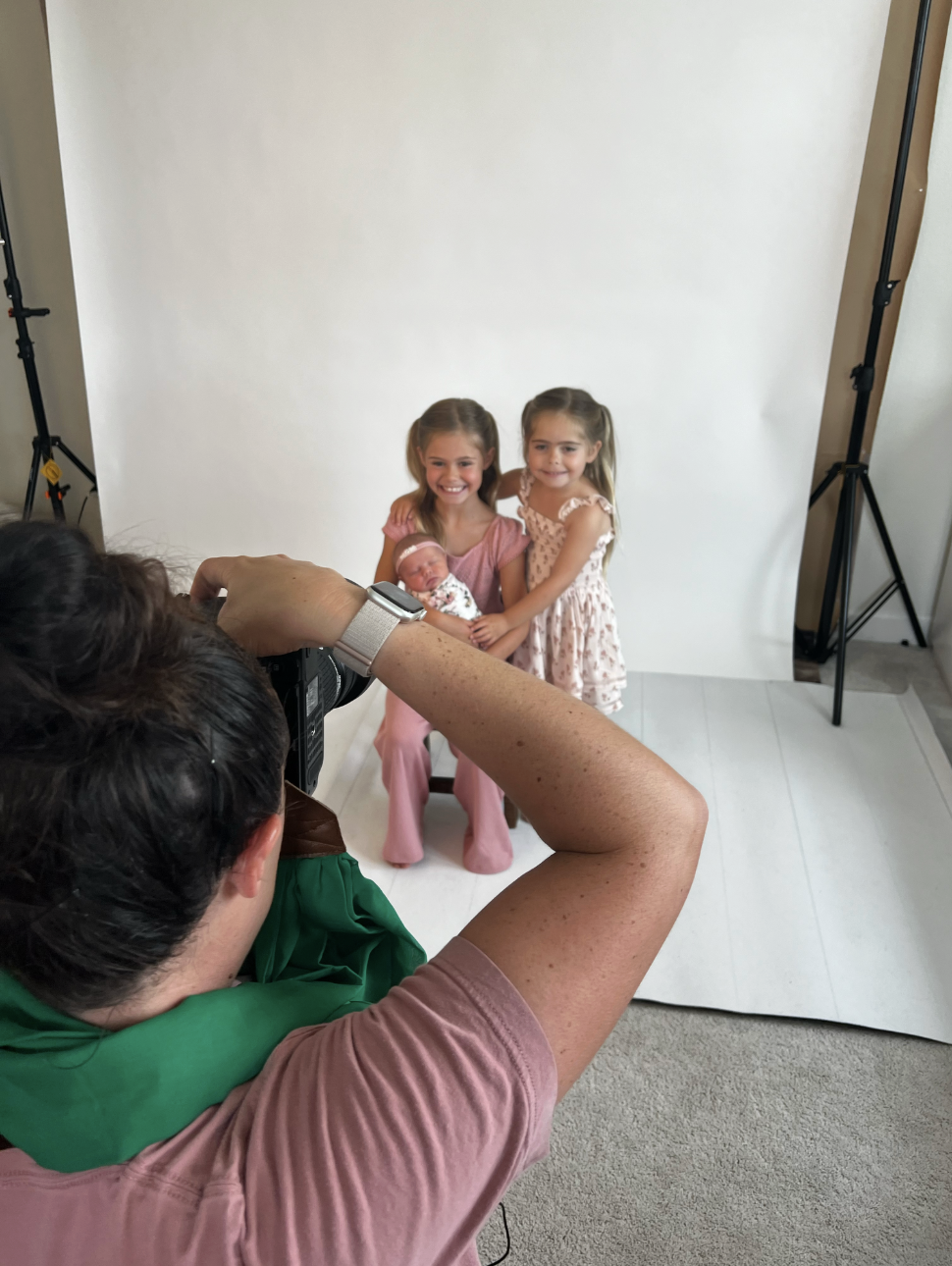Photographer taking a picture of three young girls, one holding a newborn, in a studio with a white backdrop.