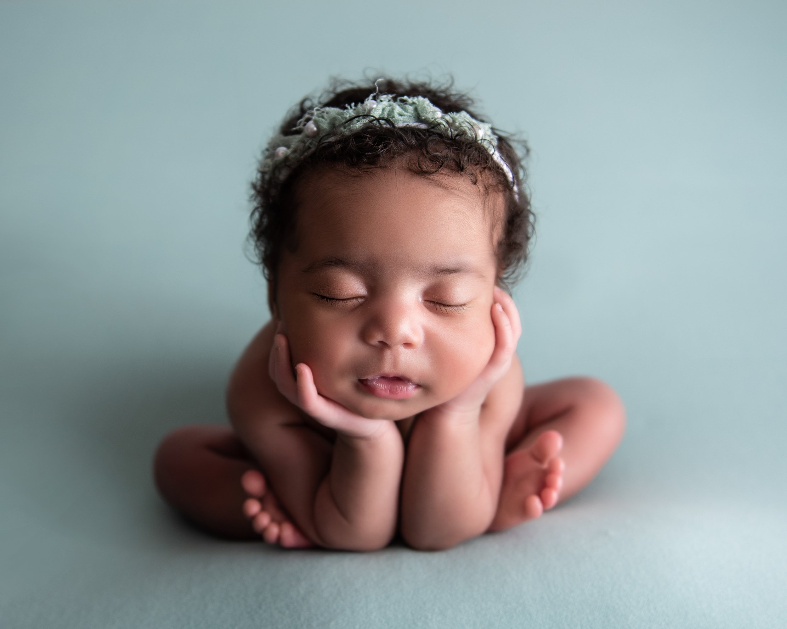 A newborn baby with curly hair and a headband, sleeping with face resting on hands against a plain blue background.