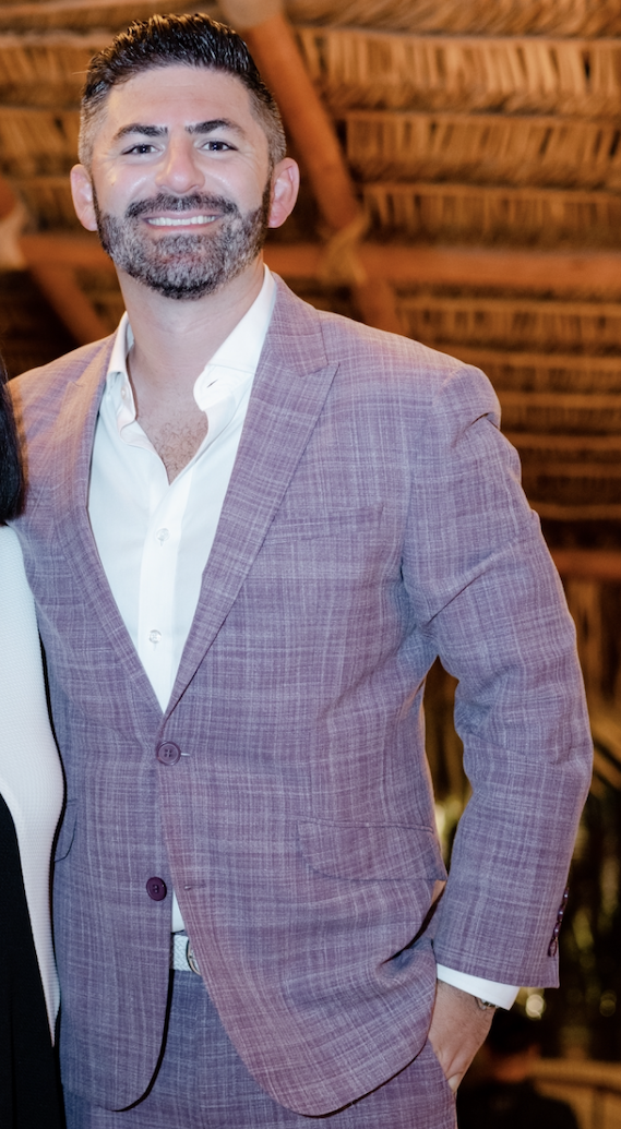 A man with dark hair and beard, wearing a light gray plaid suit jacket and white shirt, smiling at the camera indoors with wooden ceiling beams in the background.