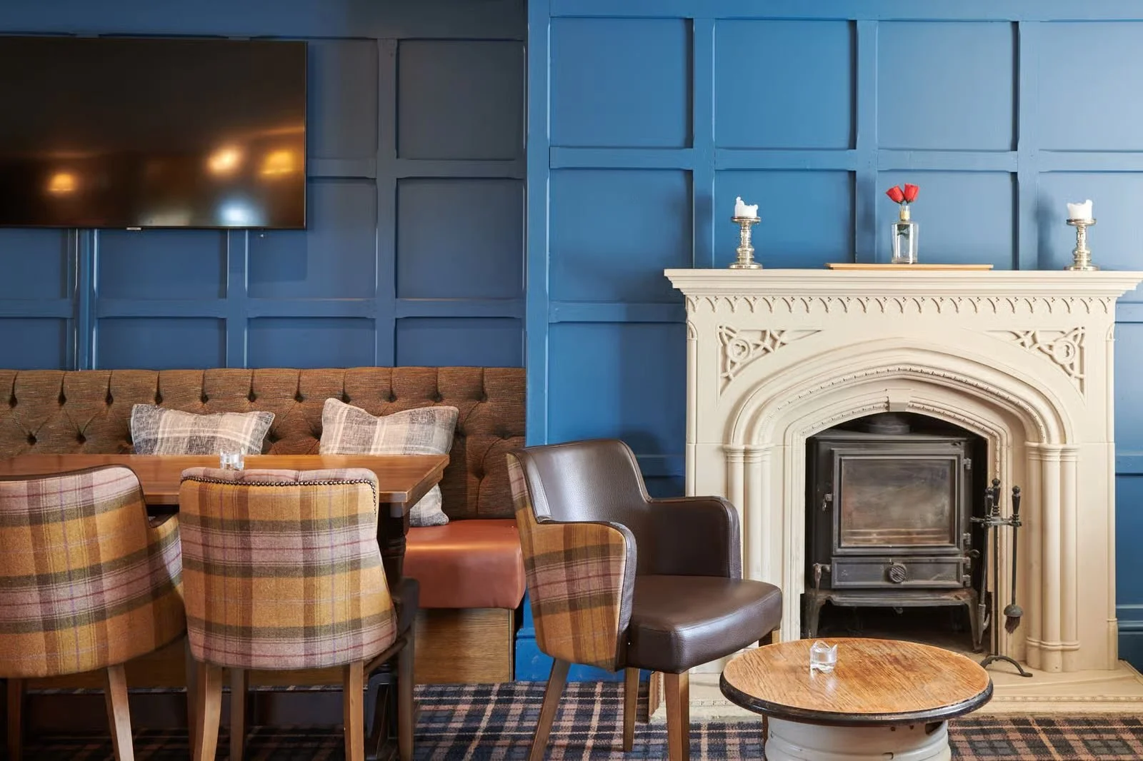Interior of a cozy room with a navy blue paneled wall, a white decorative fireplace with small candles and flowers on top, a flat-screen TV mounted on the wall, a wooden table with plaid and solid-colored chairs, a brown armchair, a small round wooden coffee table with a glass of water, and a plaid area rug.