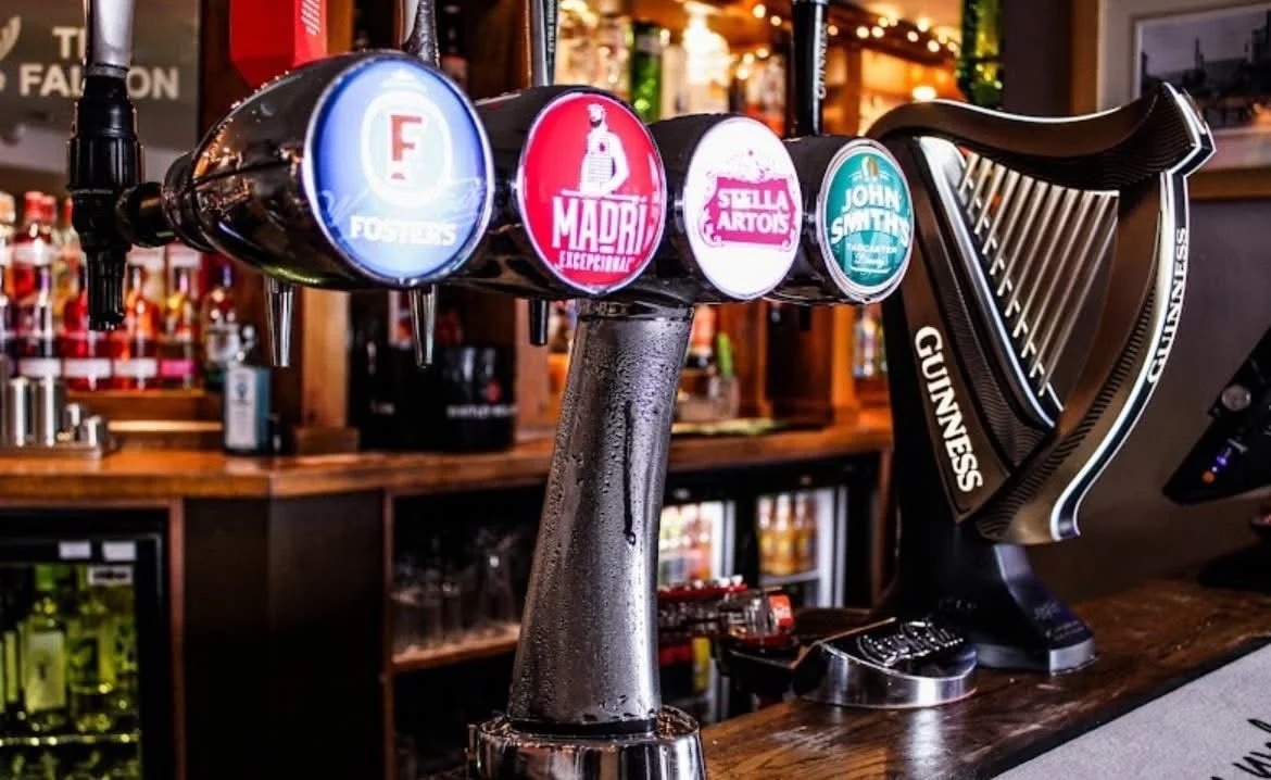 Beer tap handles with logos of Foster's, Madrid, Stella Artois, and John Smith's, next to a Guinness tap in a bar setting.