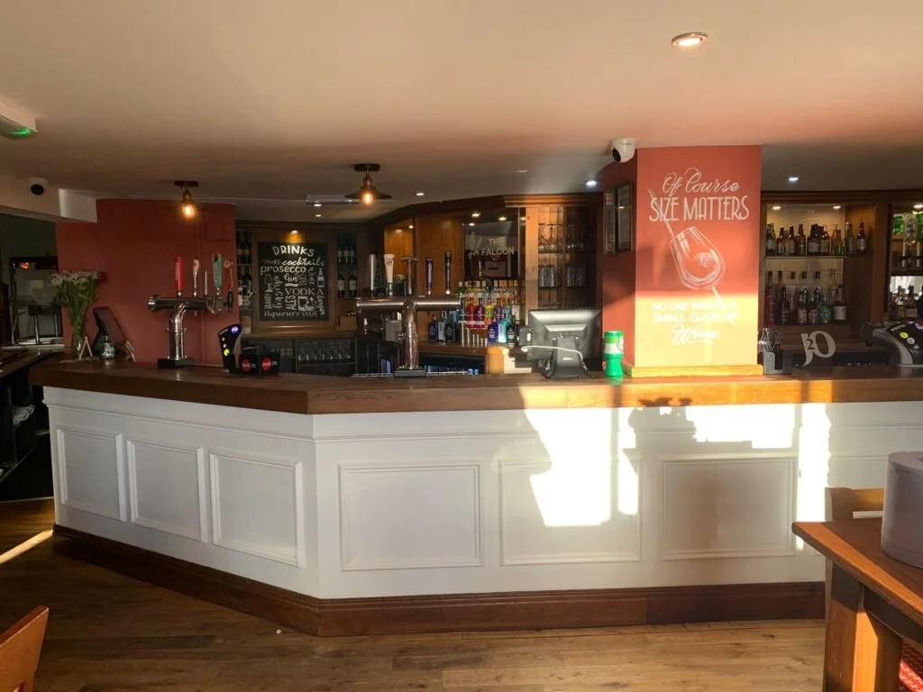 Empty bar with wooden countertop, tap handles, and a drink menu sign in the background, decorated with a red and orange wall featuring the message 'Of course, she matters' and a graphic of a wine glass.