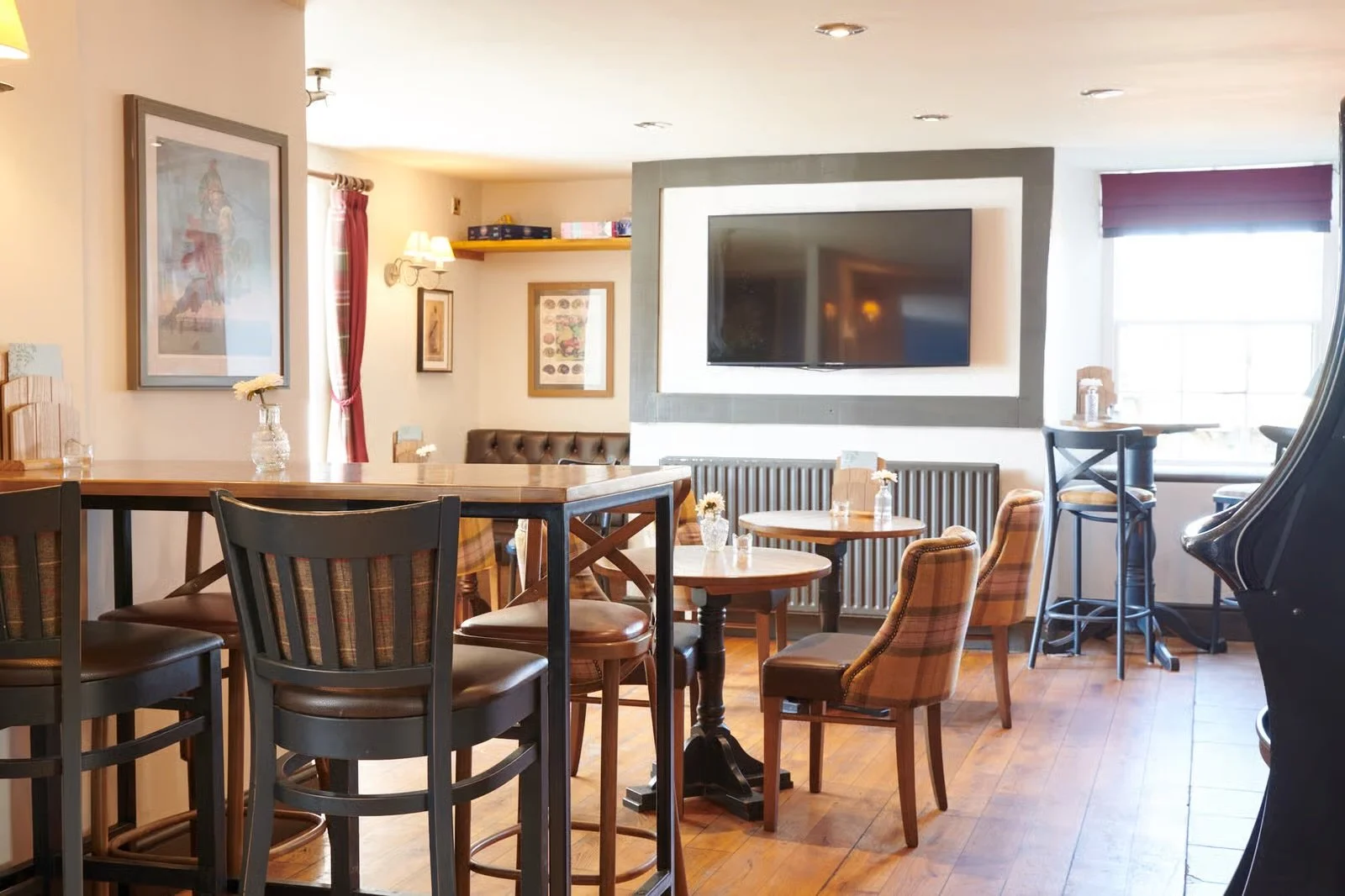 Cozy corner in a restaurant with wooden tables, mixed chairs, a large TV on the wall, framed pictures, and windows with curtains.