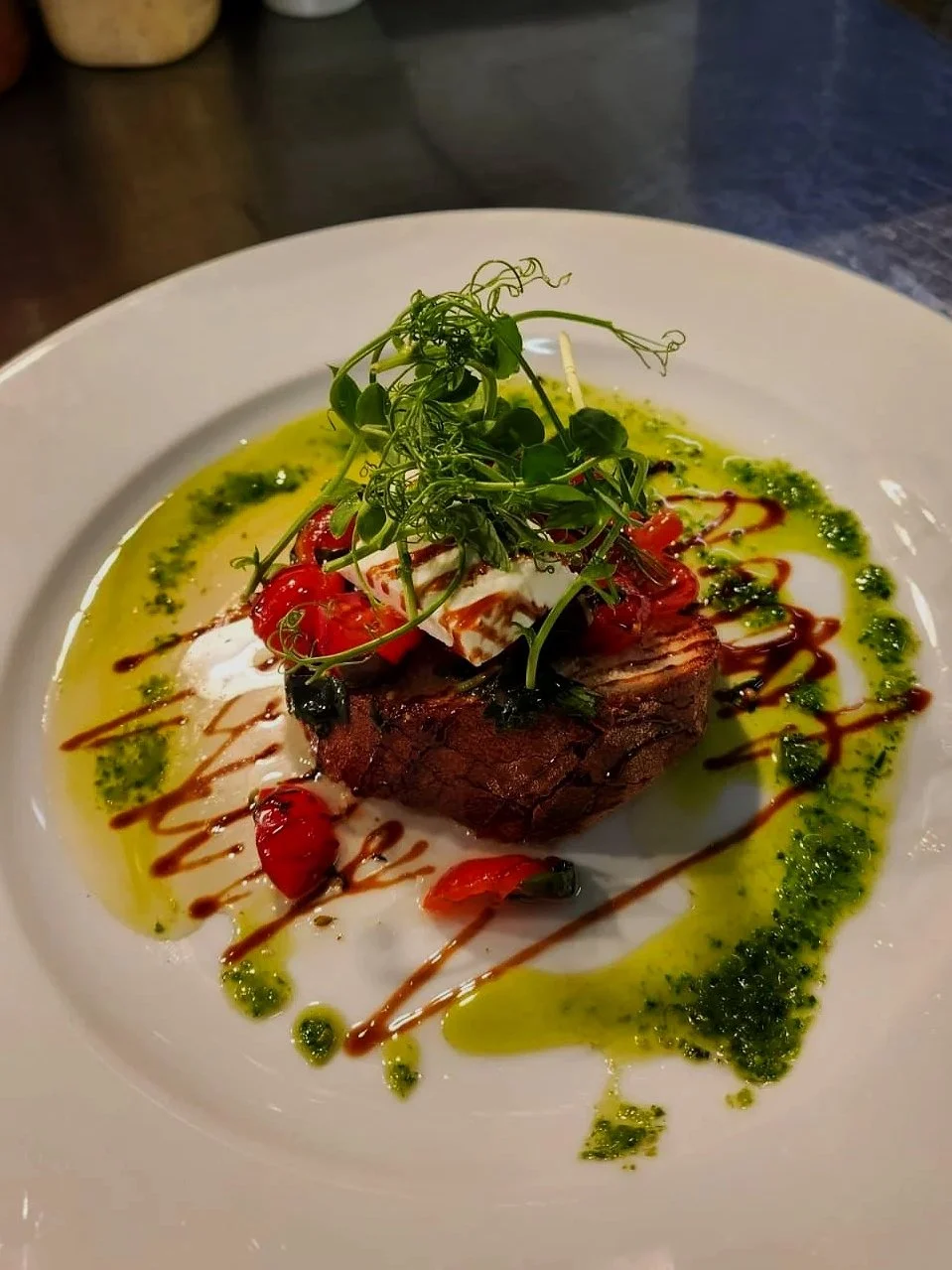 A plated dish featuring a piece of cooked beef topped with microgreens, cherry tomatoes, and drizzled with balsamic glaze. The plate is decorated with green herb oil and a white creamy sauce.