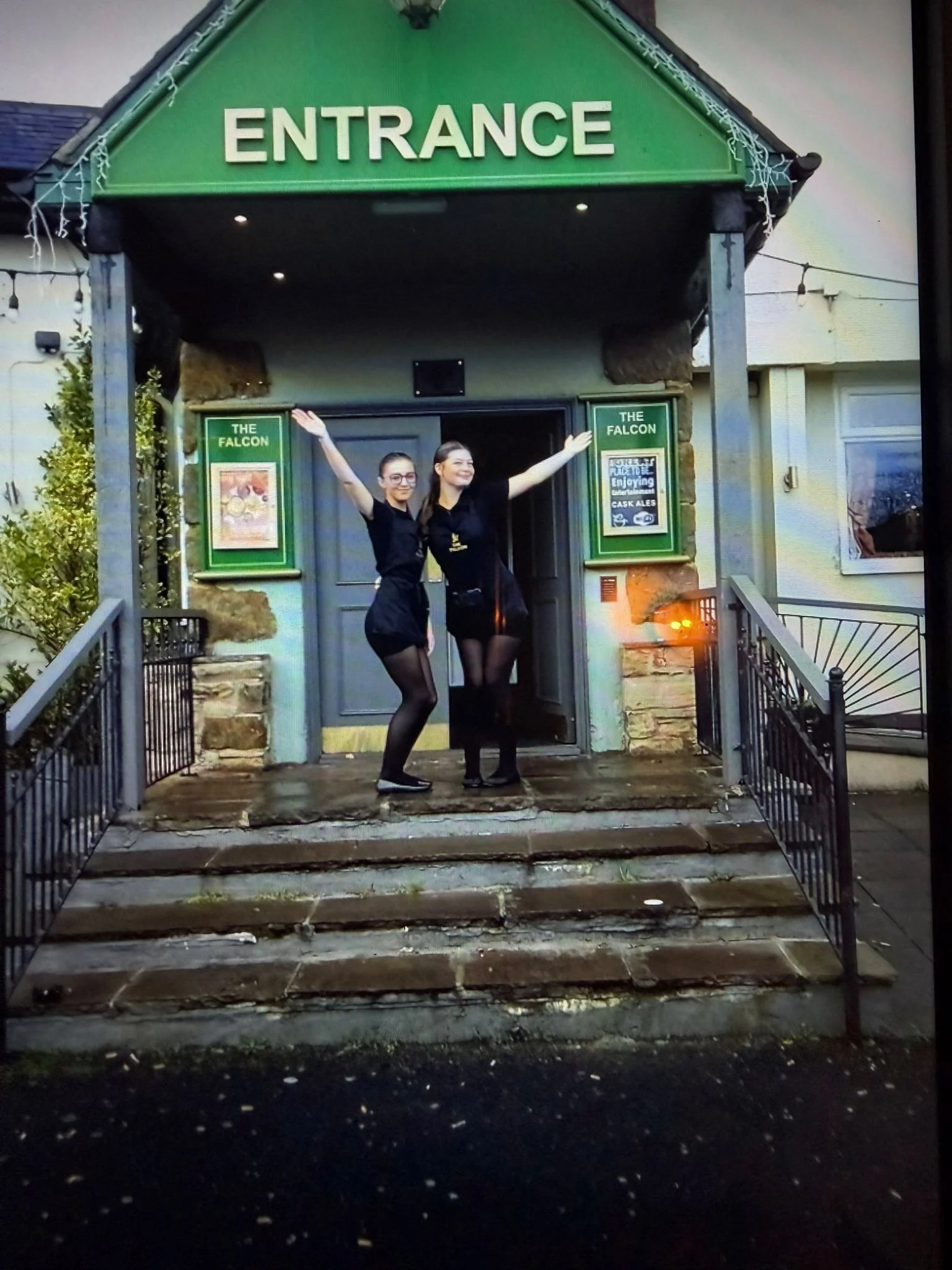 Two women in black dresses with arms raised standing on the steps of a pub called The Falcon, with a green entrance sign above them.