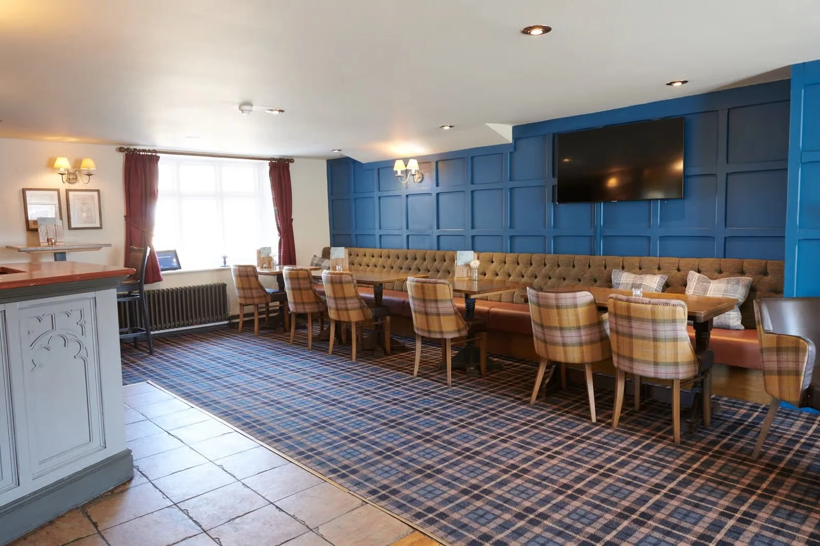 Cozy restaurant or cafe with a long plush banquette against a blue paneled wall, plaid upholstered chairs, wooden tables, a patterned carpet, a large window with red curtains, and a flat screen TV on the wall.