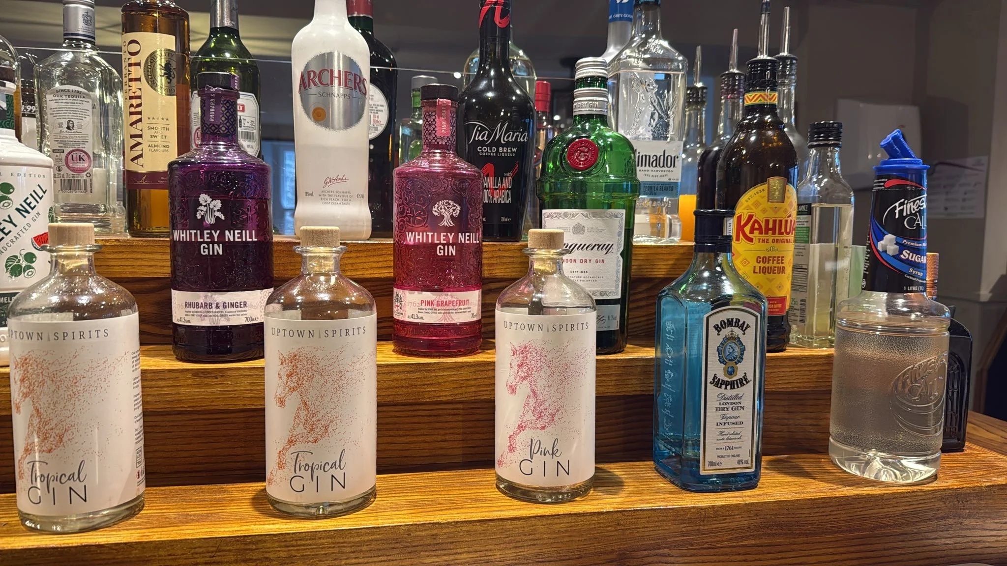 Various bottles of spirits and flavored gins displayed on a wooden bar shelf, including Whitey Neill Gin, Bombay Sapphire, Archer's Schnapps, and Kahlua.