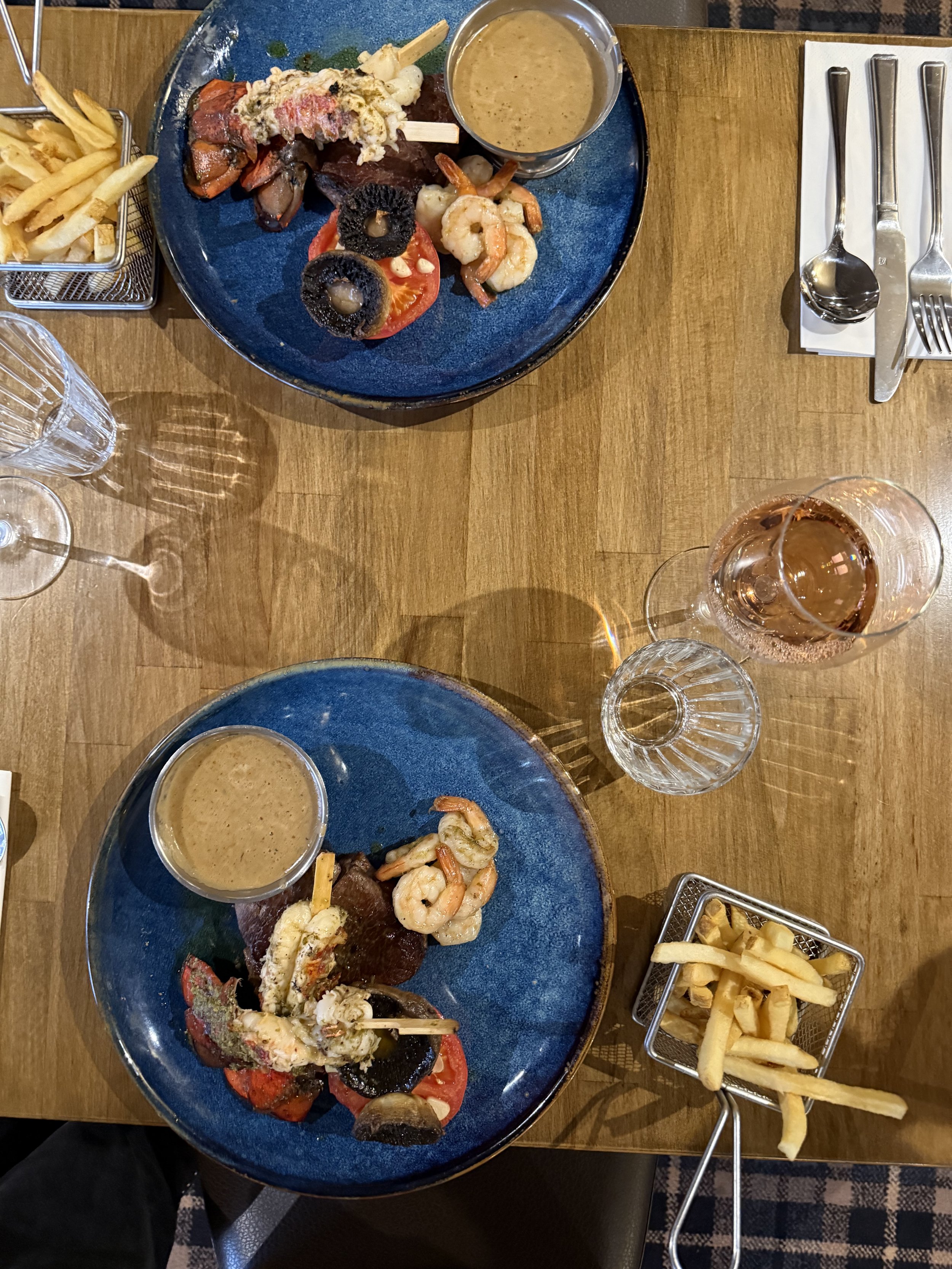 Two blue plates with seafood dishes, including shrimp, cherry tomatoes, and grilled vegetables, accompanied by a small bowl of dipping sauce on each plate. There are side views of French fries in metal baskets, drinking glasses, and silverware on a wooden table.
