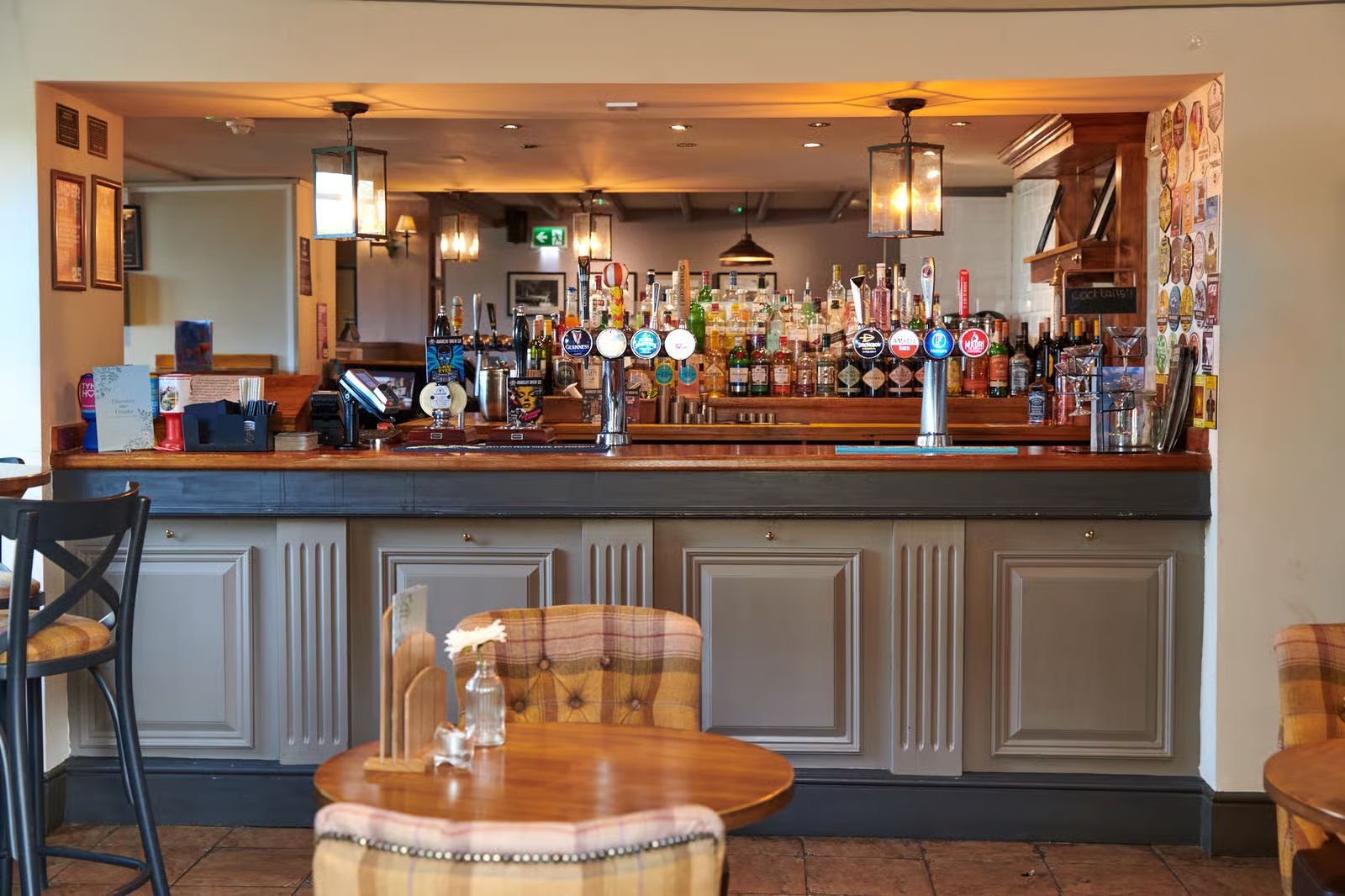 Bar counter inside a restaurant or pub with various alcohol bottles and beer taps, seating area with tables and chairs, warm lighting, and decorative wall art.