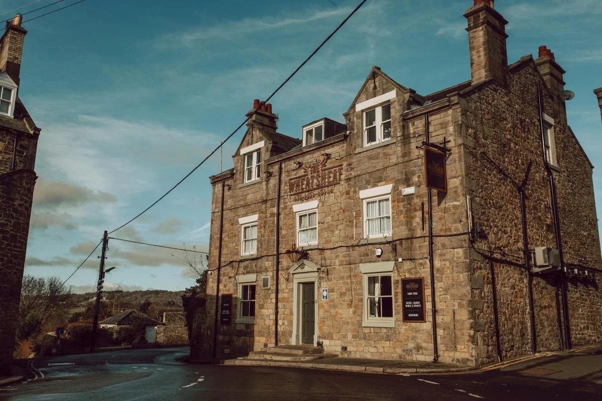 A historic stone building on a street corner with signs indicating it is a pub or inn, surrounded by a rural landscape and a partly cloudy sky.