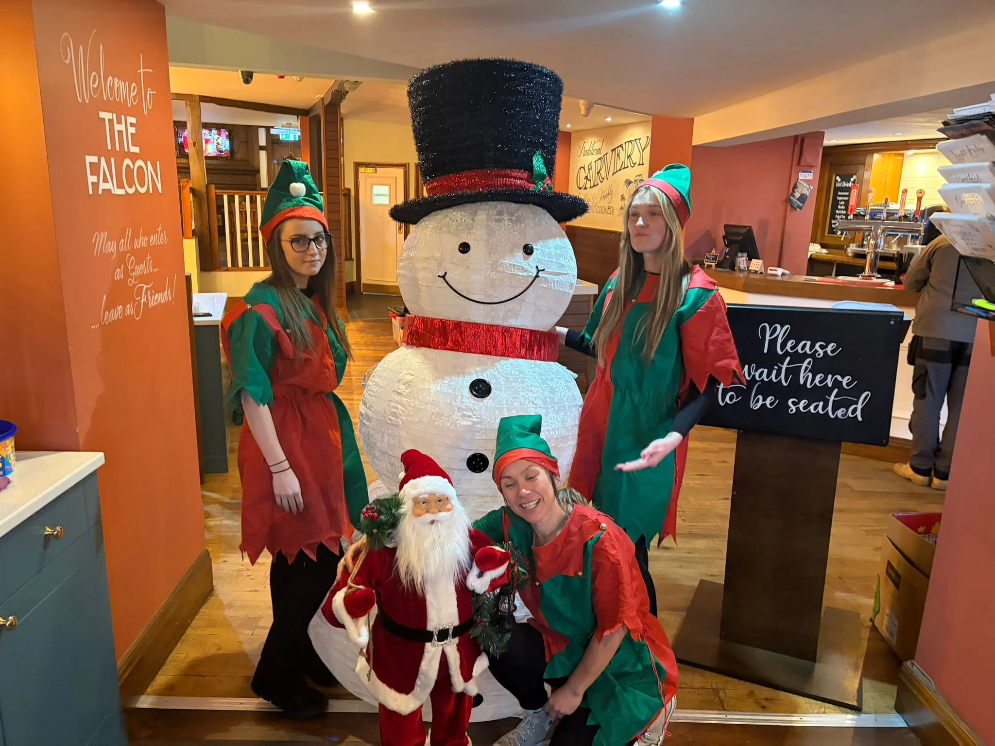 Three people dressed as elves and a Santa figure posing with a large snowman decoration inside a restaurant or cafe.