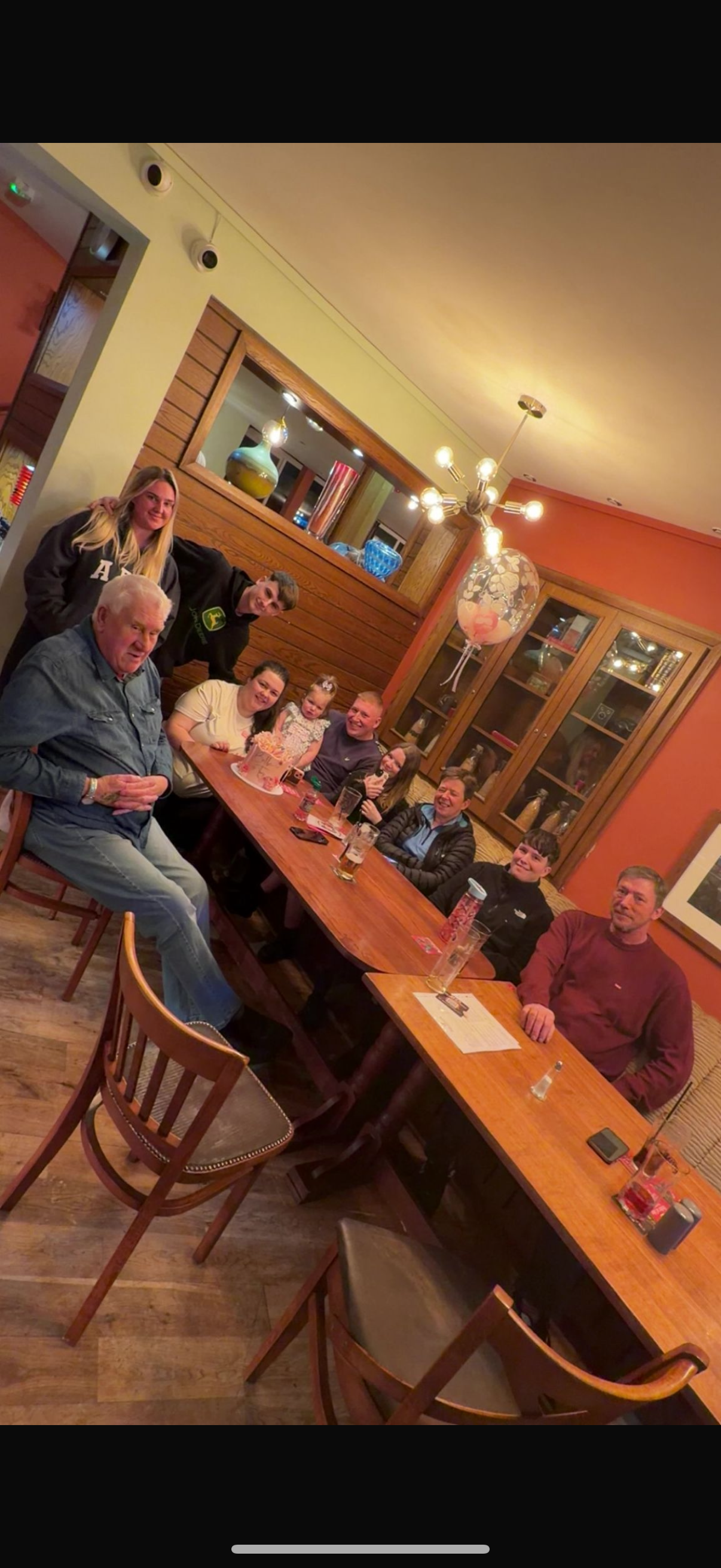A group of people celebrating around a long wooden table at a restaurant, with drinks and a cake, some with smiles and laughter, under warm lighting and colorful decor.
