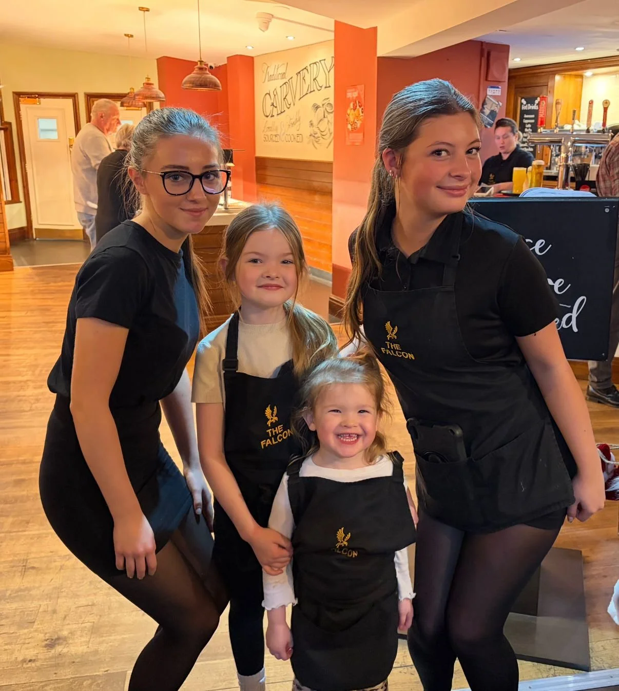 Four young girls wearing black aprons labeled "The Falcon" smiling and posing inside a restaurant or café with warm lighting and wooden decor.