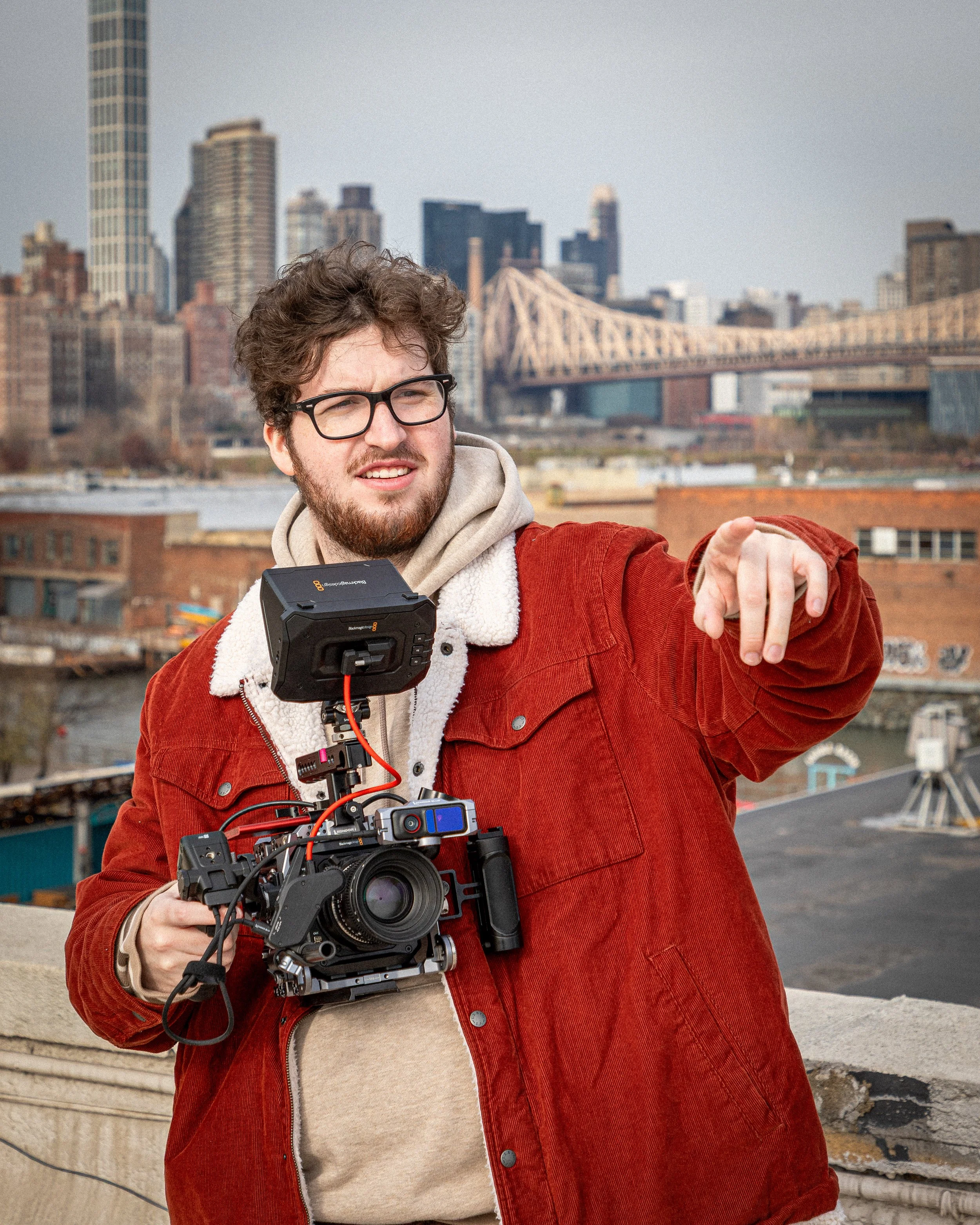 A young man with curly hair and glasses pointing with his right hand on a rooftop during the daytime, with a city skyline including bridges and skyscrapers in the background, while holding a professional camera rig.