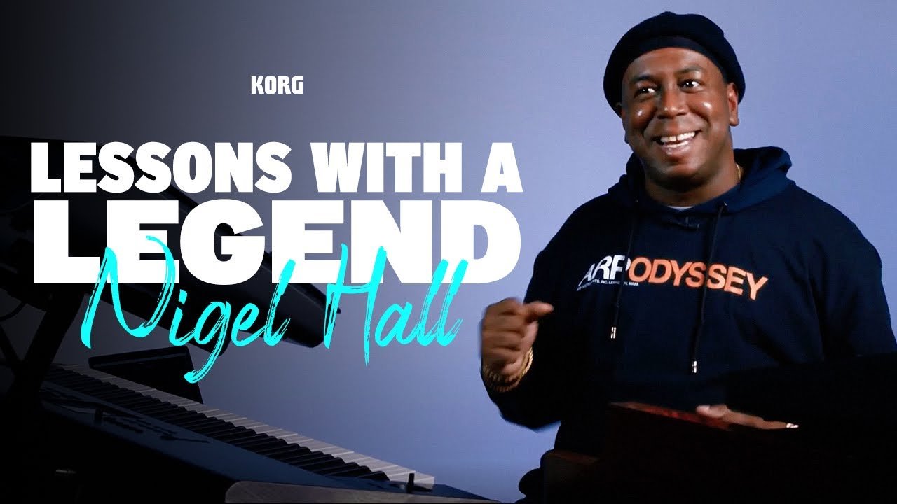 A man smiling and wearing a black beanie and hoodie, holding a tablet, standing next to a black grand piano. The background is blue-gray, with text that reads 'Lessons with a Legend Nigel Hall,' and the Korg logo is visible.