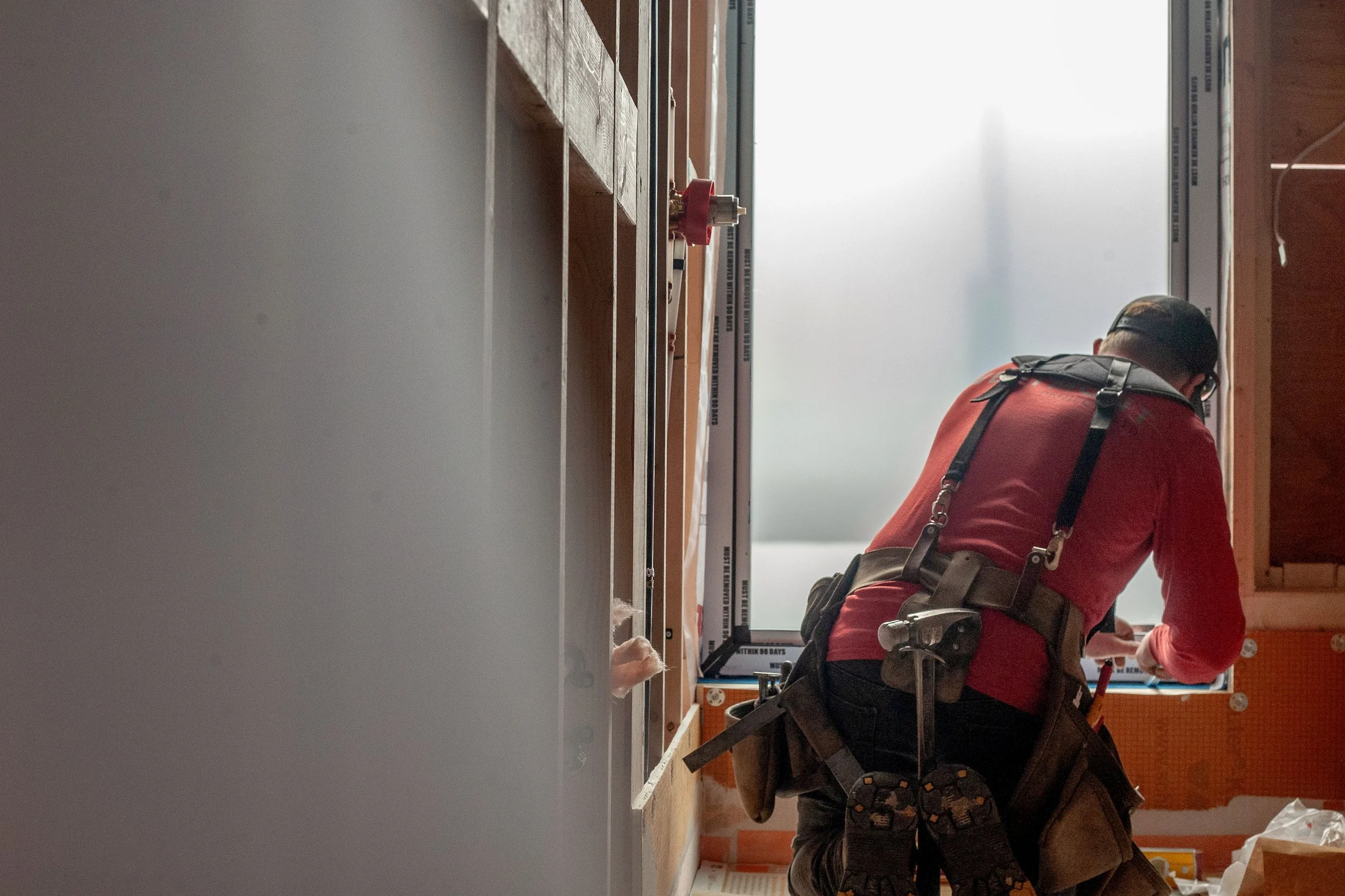 A construction worker installing a window in a building, seen from behind, with the worker wearing a red shirt and a tool belt, working on the window frame near an unfinished wall.