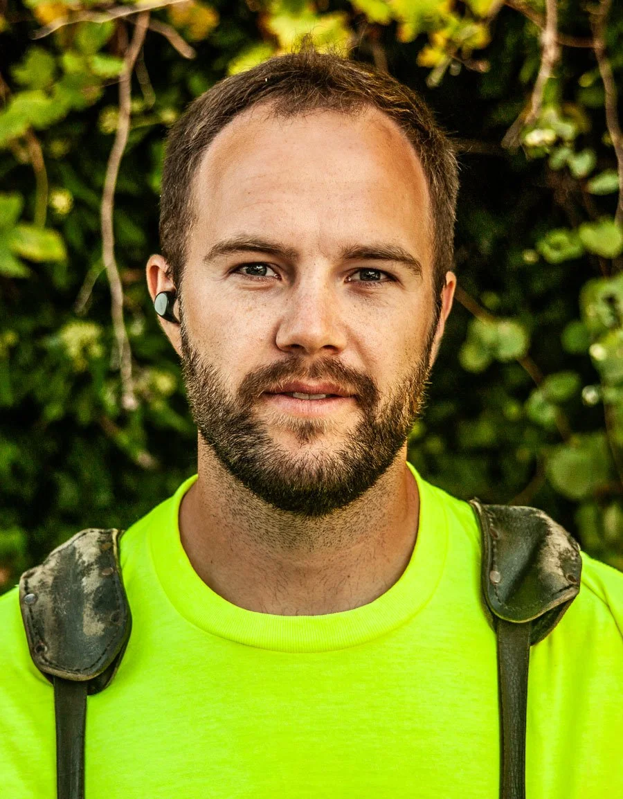A young man with a beard wearing a bright yellow shirt and a backpack standing outdoors in front of green foliage.