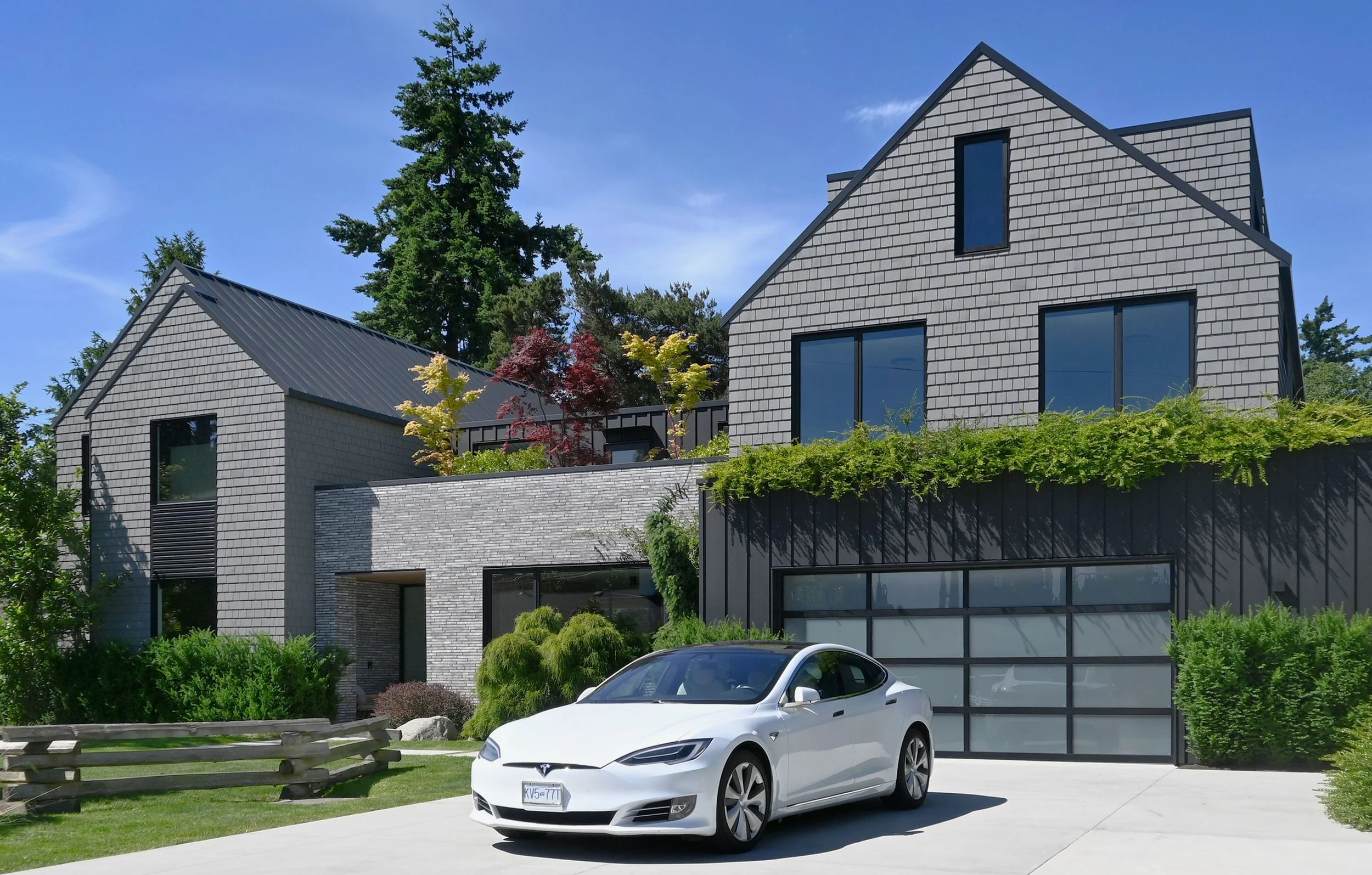 Modern house with grey brick and siding, large windows, green plants, and a white Tesla parked in driveway.