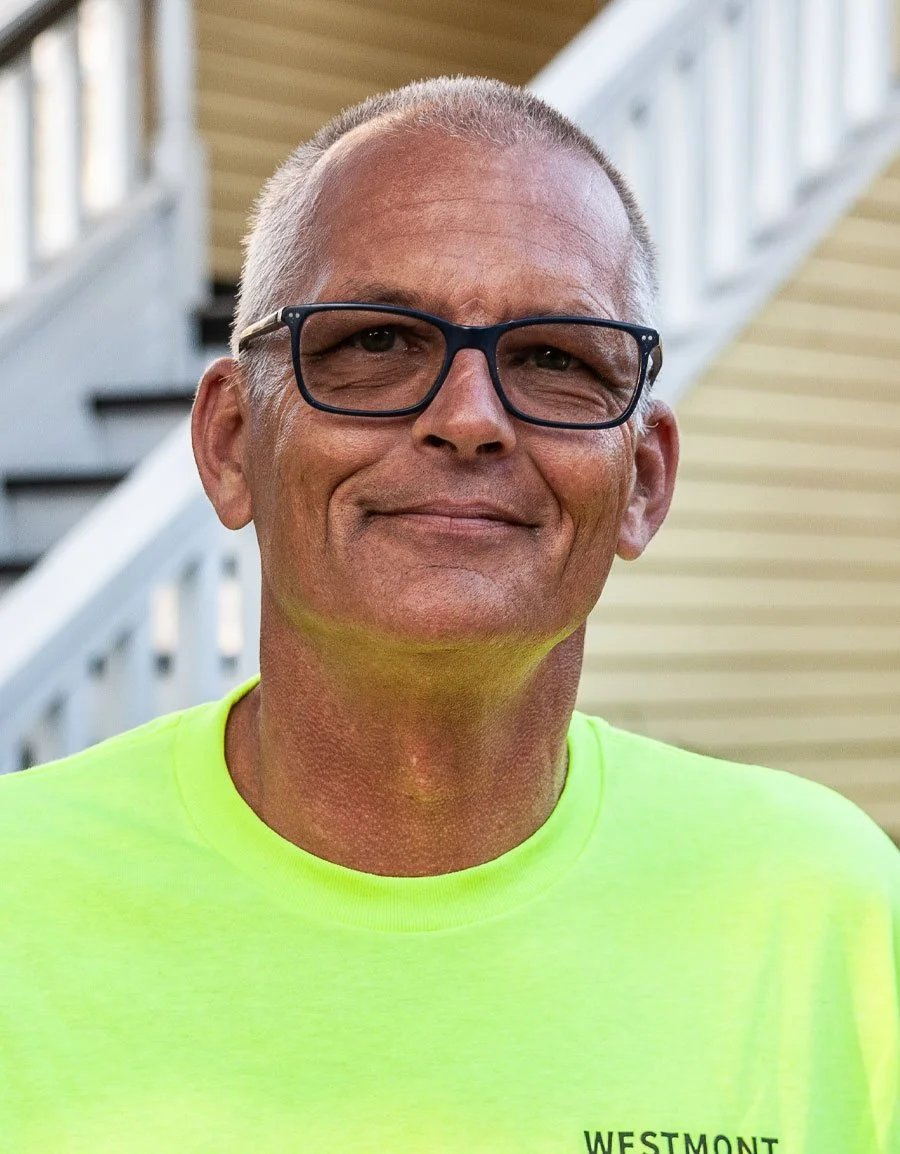 A smiling middle-aged man with short gray hair, wearing black-framed glasses and a bright yellow T-shirt, standing outdoors near a staircase.