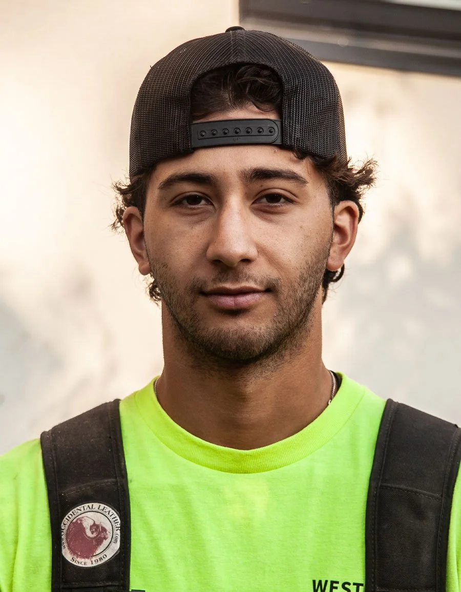 A young man wearing a black trucker hat backwards, a neon yellow shirt, and a backpack, standing outdoors against a plain background.