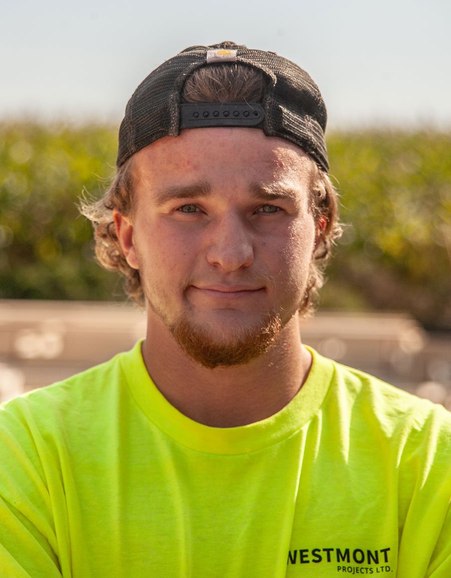A young man with light brown curly hair and a beard, wearing a black backward baseball cap and a bright yellow shirt with black text, standing outdoors with a blurred background of greenery and a clear sky.