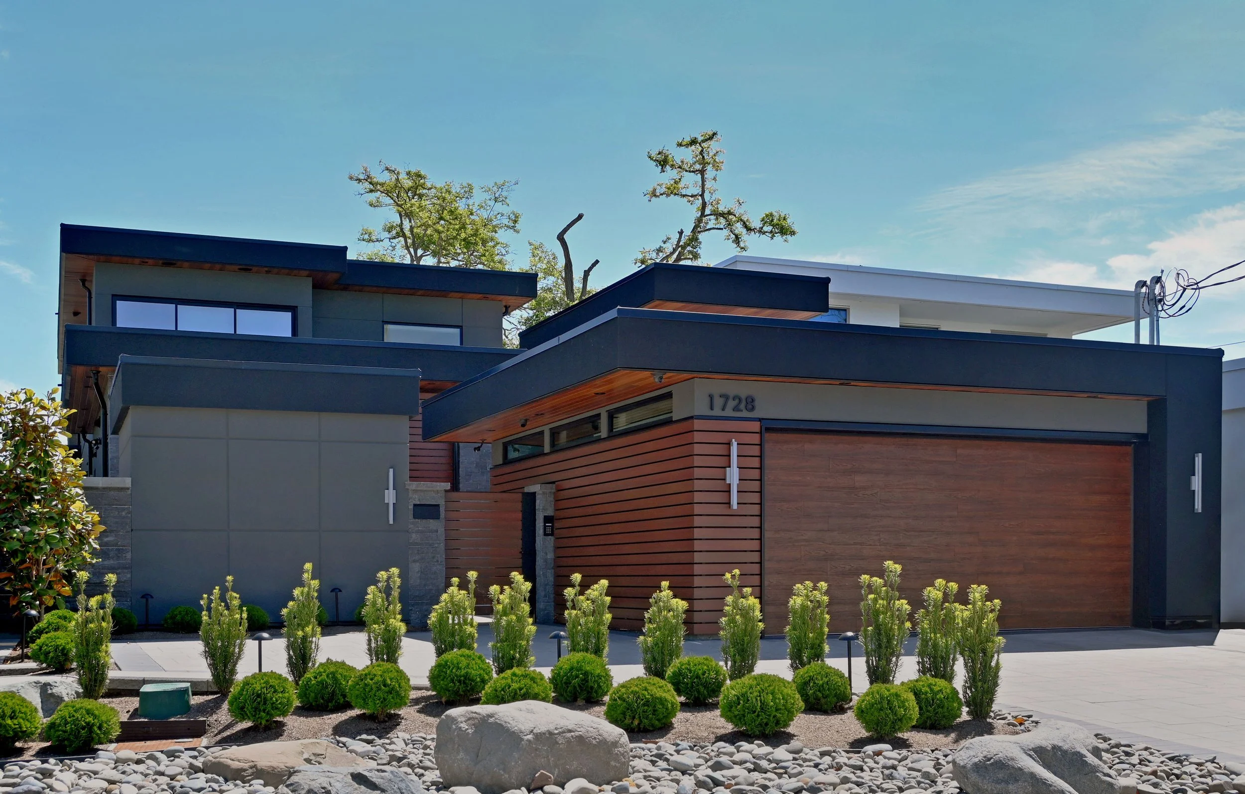 Modern house with a combination of wood, concrete, and black metal exterior. Front yard has lush green bushes, small trees, and decorative rocks. Clear blue sky in the background.