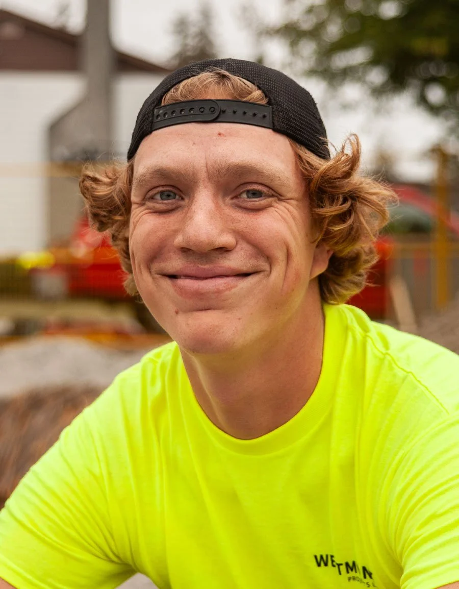 A young man with curly red hair, wearing a black backward baseball cap and a bright yellow T-shirt, smiling outdoors with blurred trees and a red structure in the background.