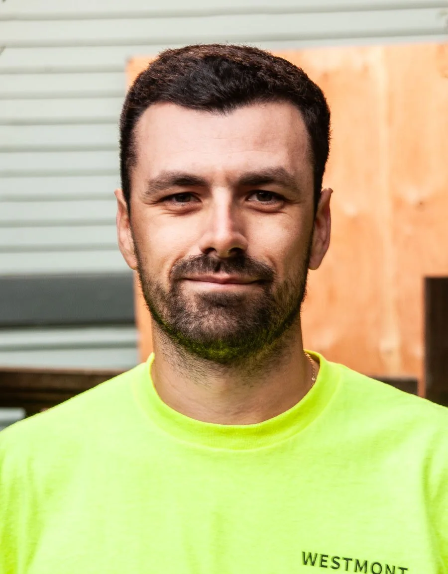 A man with dark hair and a beard wearing a bright yellow shirt, standing indoors with a wooden wall and window behind him.