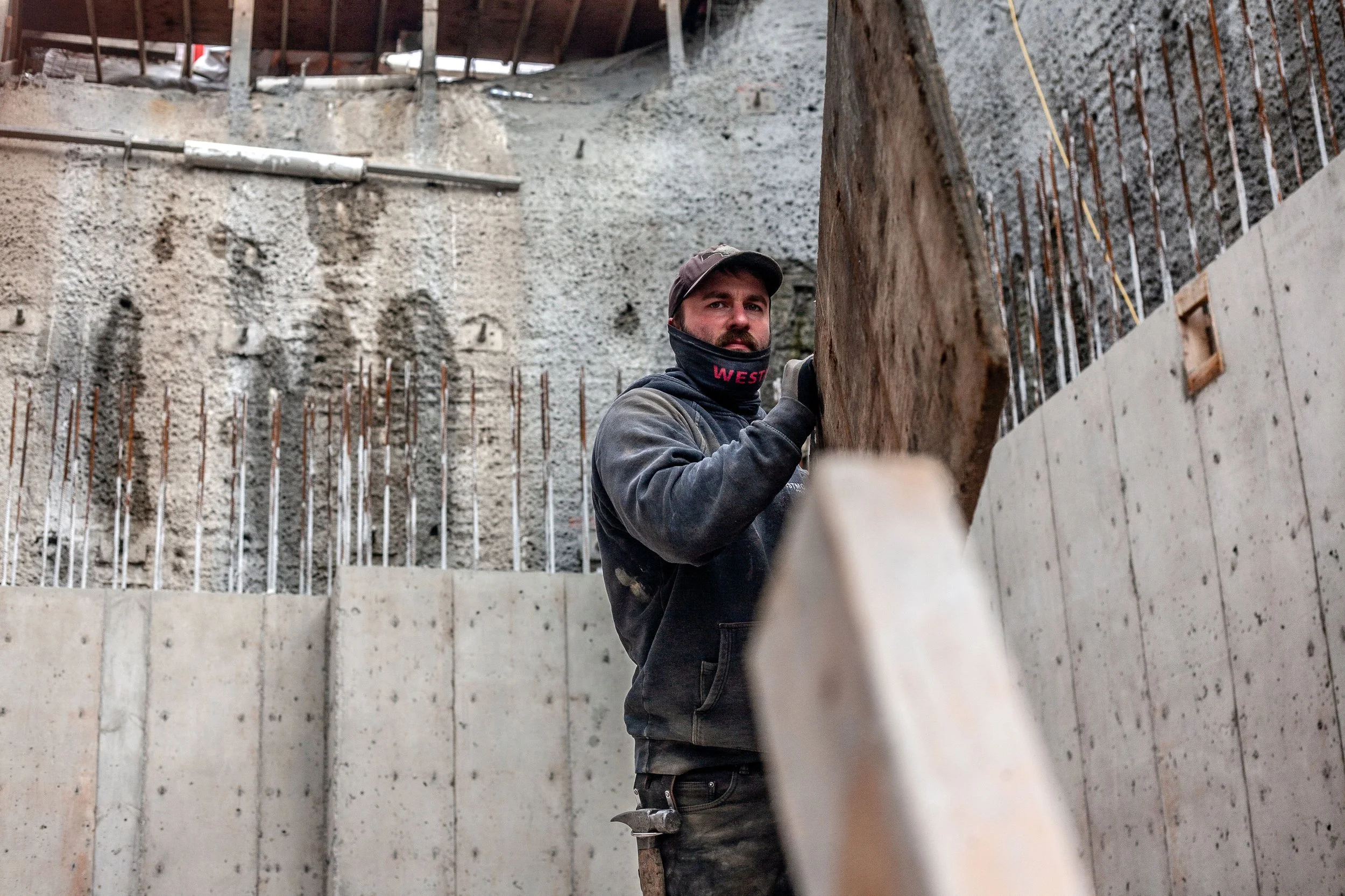 A construction worker wearing a hoodie, face mask, and gloves standing inside a building under construction, holding a wooden plank.