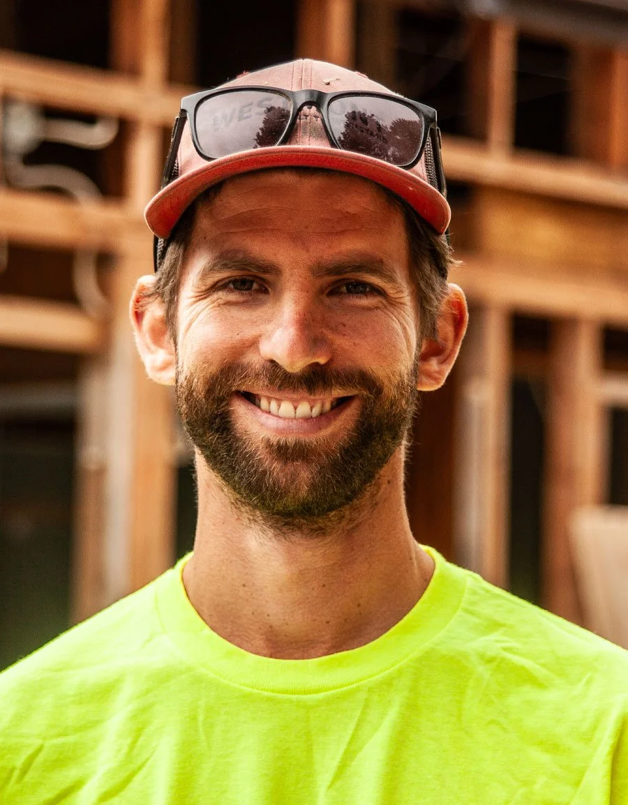 A smiling man with a beard and mustache wearing a neon yellow shirt, a red cap, and sunglasses on top of the cap, standing outdoors with wooden structures in the background.