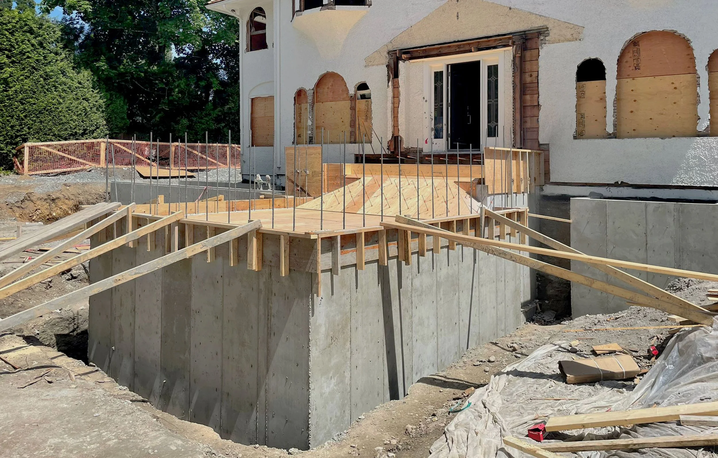 Construction site showing a house under renovation with wooden framing and concrete foundation, with some windows boarded up, and construction materials scattered around.