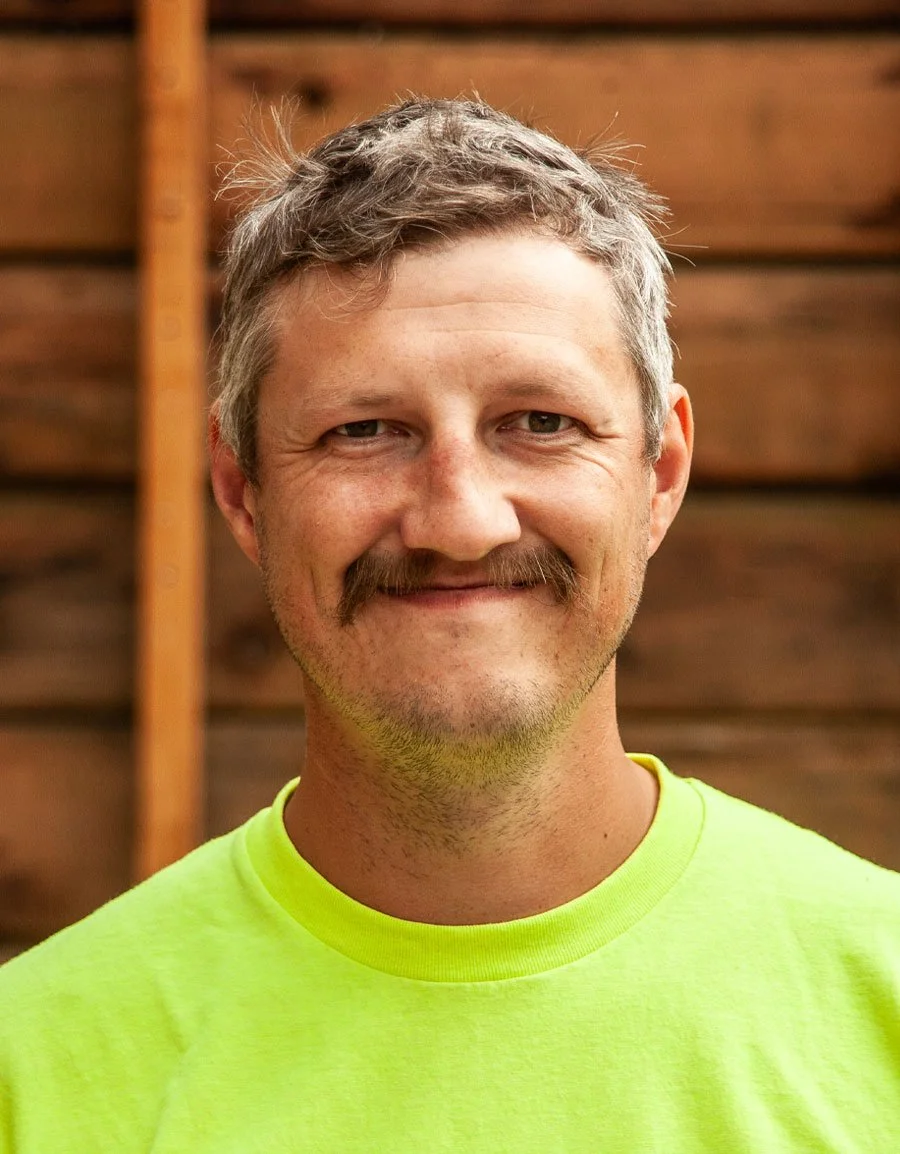 A man with gray hair and a mustache wearing a bright yellow T-shirt, smiling, standing outdoors in front of a wooden fence.