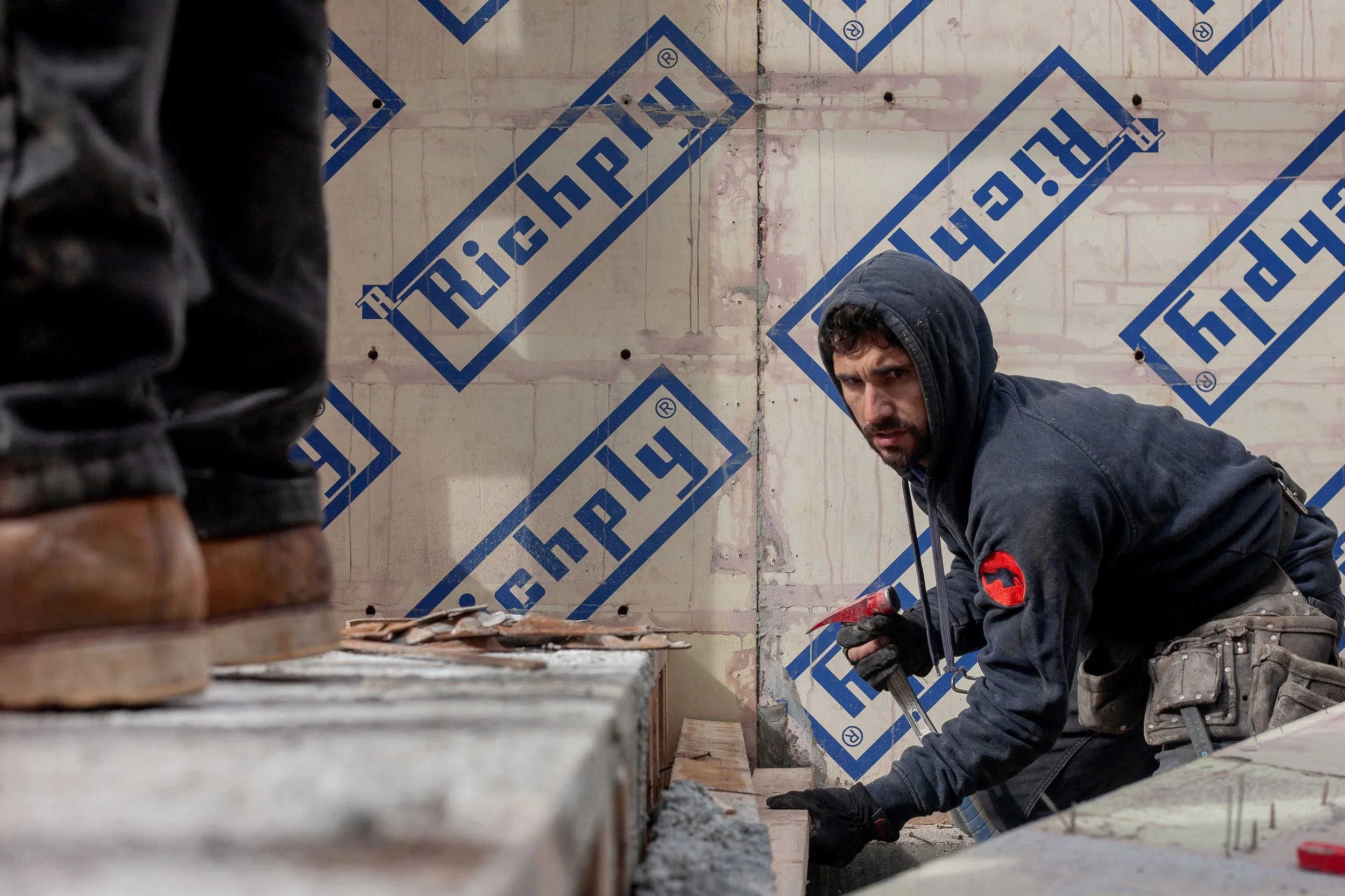 Construction worker in a hoodie crouching on a construction site, working with tools on a concrete surface, with unfinished walls marked with blue 'Richply' brand on construction sheathing.