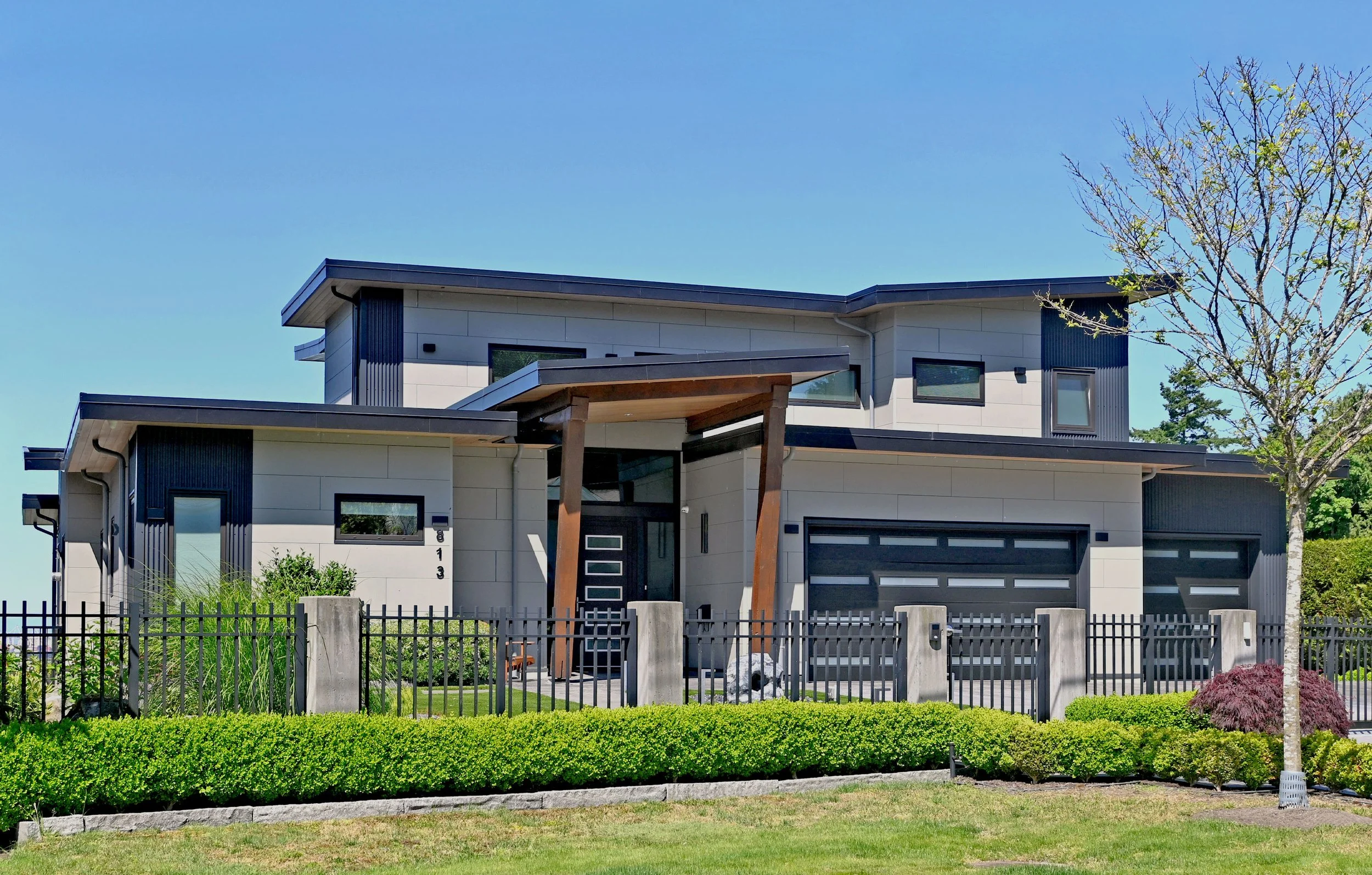 Modern two-story house with black and white exterior, large garage doors, wooden columns at the entrance, surrounded by a black metal fence and green landscaped yard with bushes and a tree.