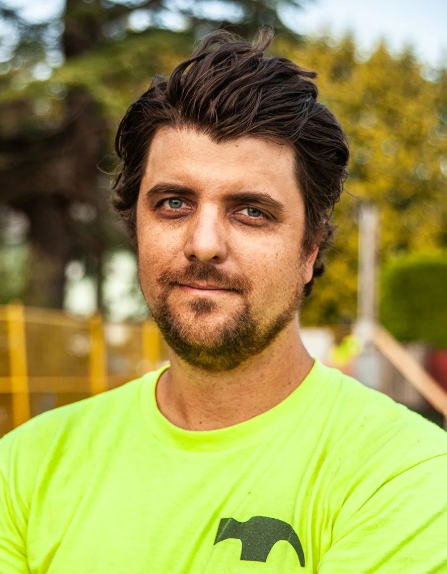 A man with dark brown hair and piercing blue eyes stands outdoors wearing a bright yellow shirt with a black logo. The background features trees and fencing, suggesting a park or recreational area.