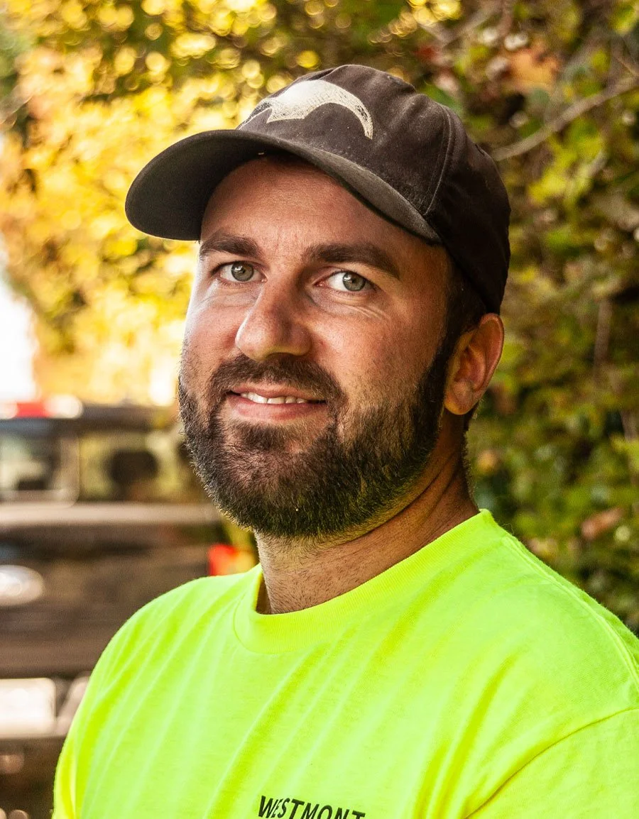 A man with a beard wearing a black cap and a neon yellow shirt, standing outdoors with autumn-colored trees in the background.