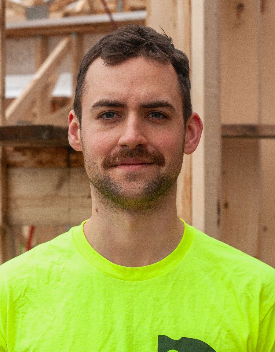 A young man with short brown hair and a mustache wearing a bright yellow shirt standing indoors with unfinished wooden walls and construction materials in the background.