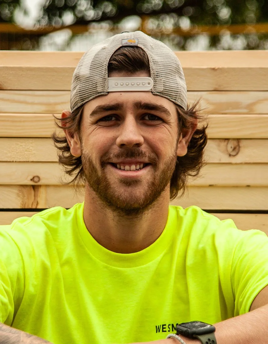 A smiling man wearing a light-colored trucker hat, a bright yellow t-shirt, and a black wristwatch, sitting outdoors in front of a wooden fence.