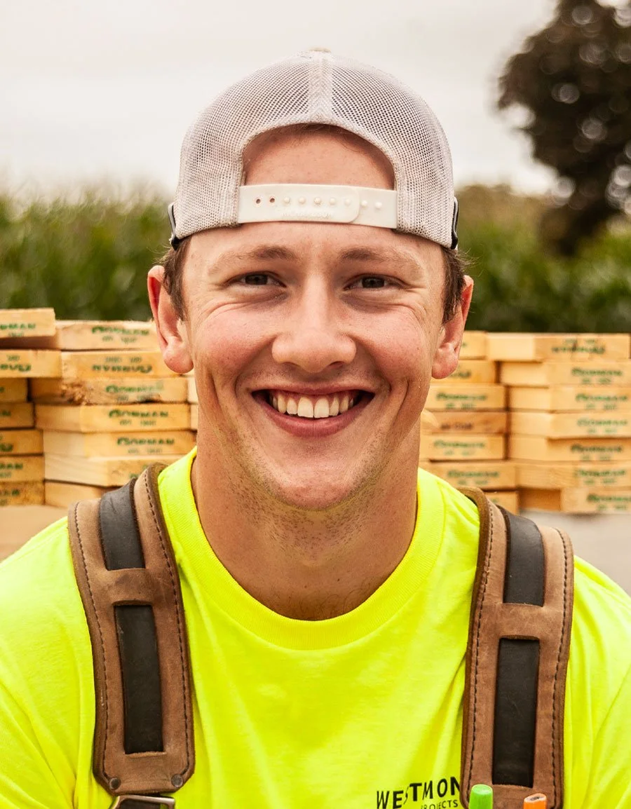 A young man with a smiling face, wearing a gray and white trucker hat backwards, a bright yellow shirt, and a brown backpack, standing outdoors with stacks of wood behind him.