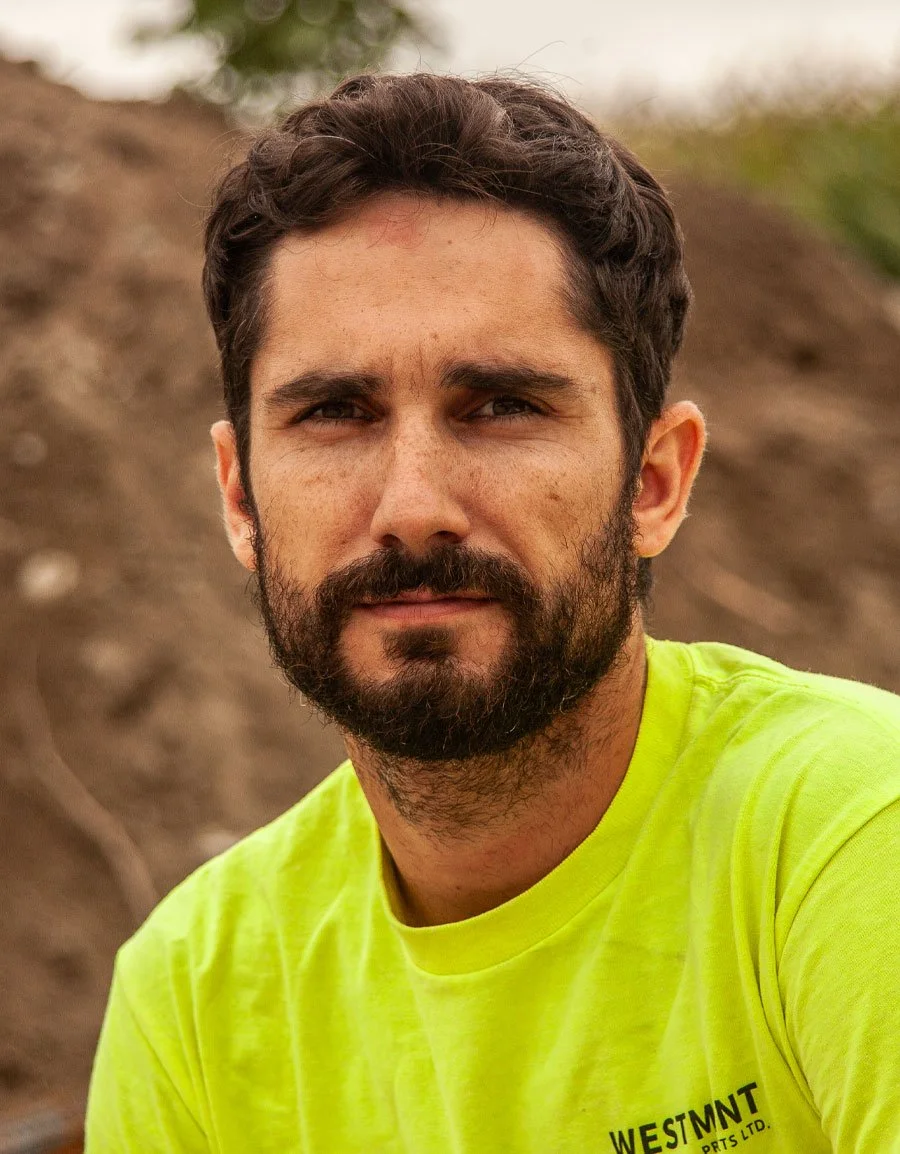 A man with dark hair and a beard, wearing a bright yellow shirt, looking at the camera with a serious expression outdoors. Background appears to be a dirt surface and some greenery.