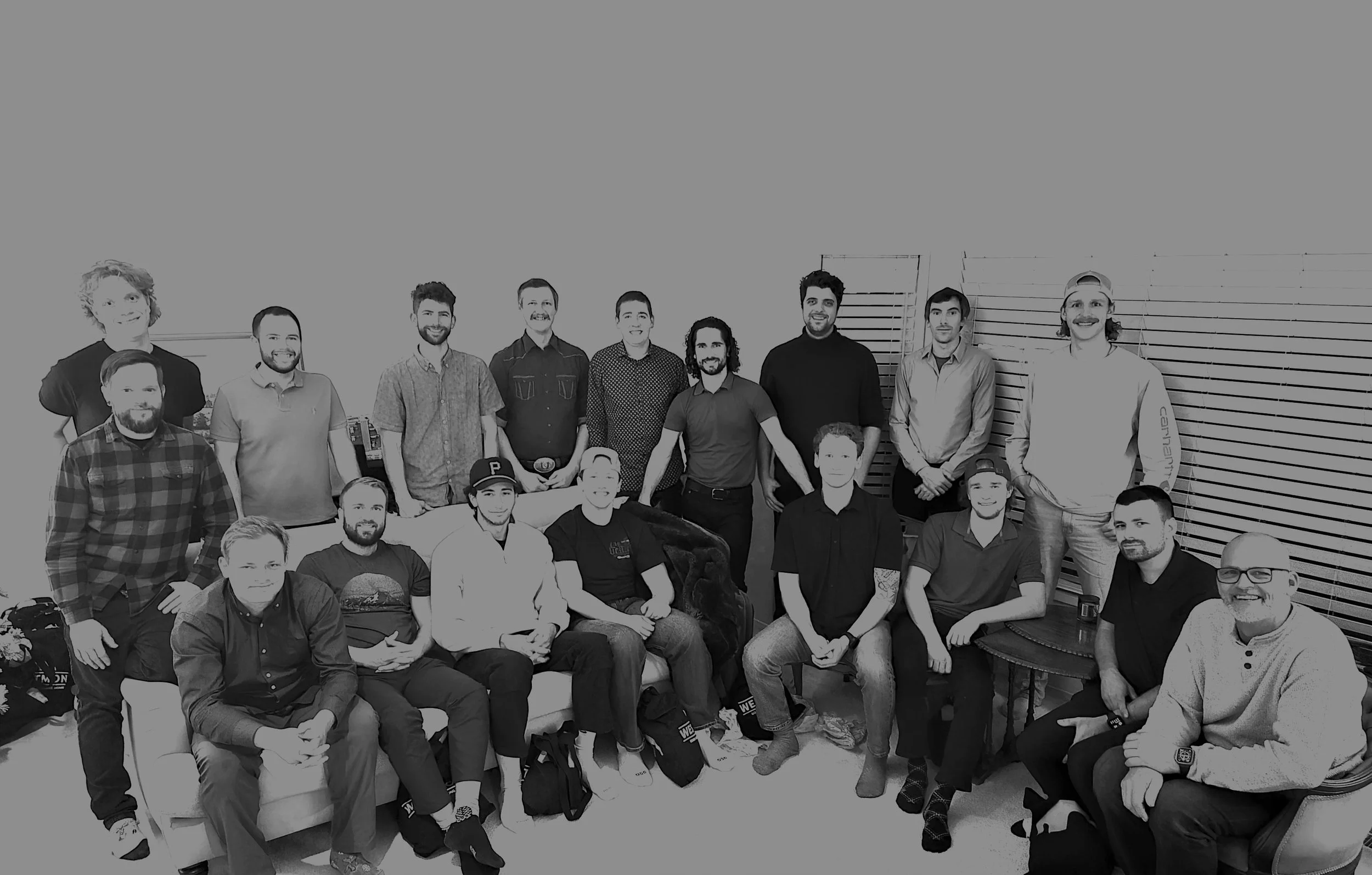 Group of 16 people in an indoor setting, some seated and others standing, smiling for a photo. The background features a plain wall and window blinds.