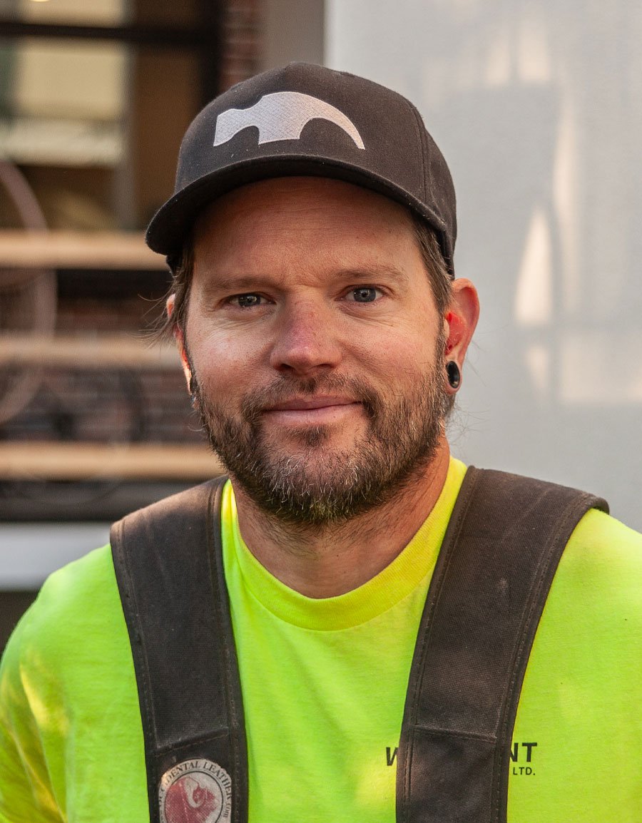 A man with a beard wearing a black cap, black earrings, and a bright yellow shirt, standing outdoors with a blurred background.