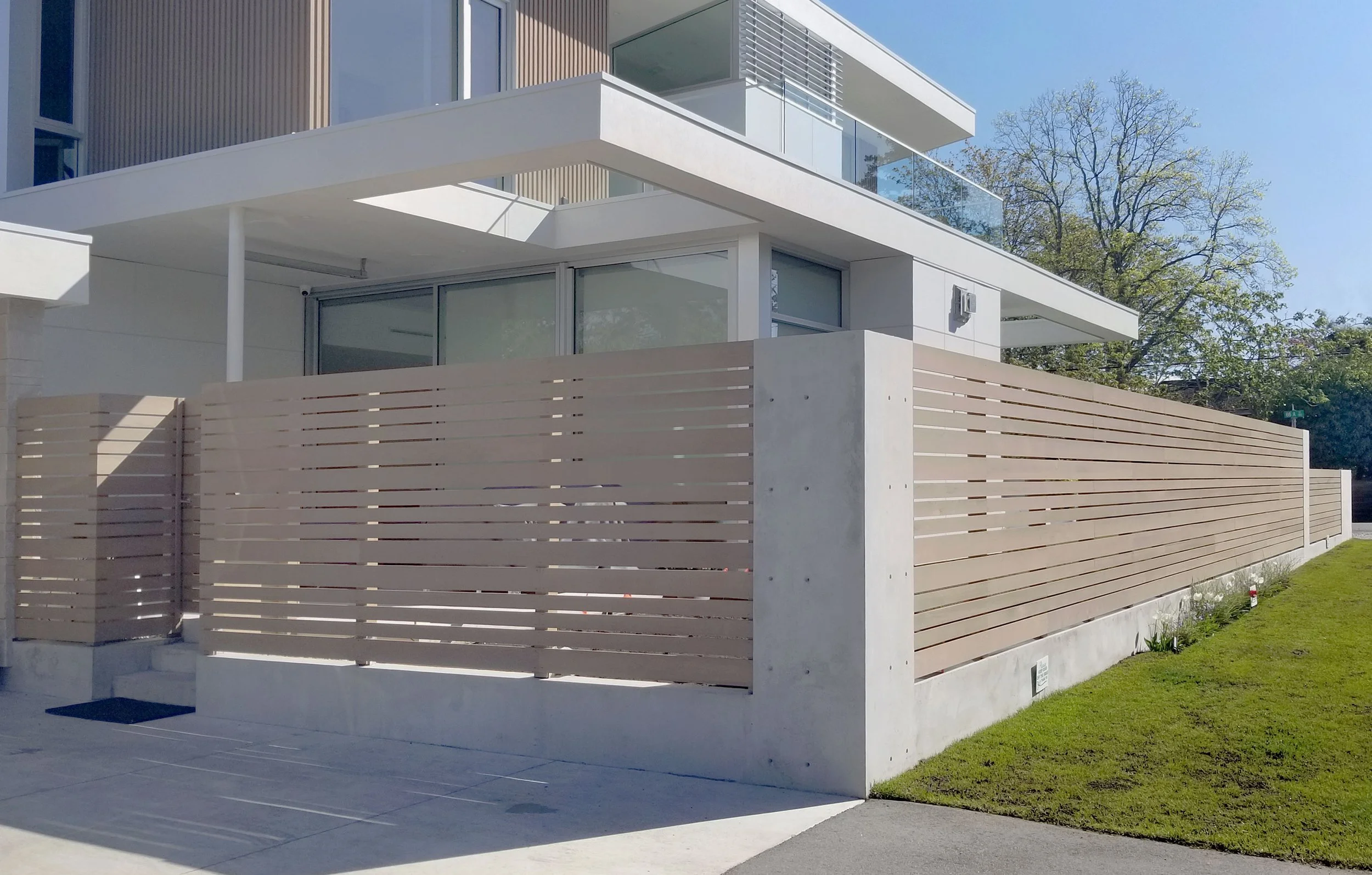 Modern residential building with a beige horizontal wooden fence, large glass windows, white walls, and a well-kept lawn, under a clear blue sky.