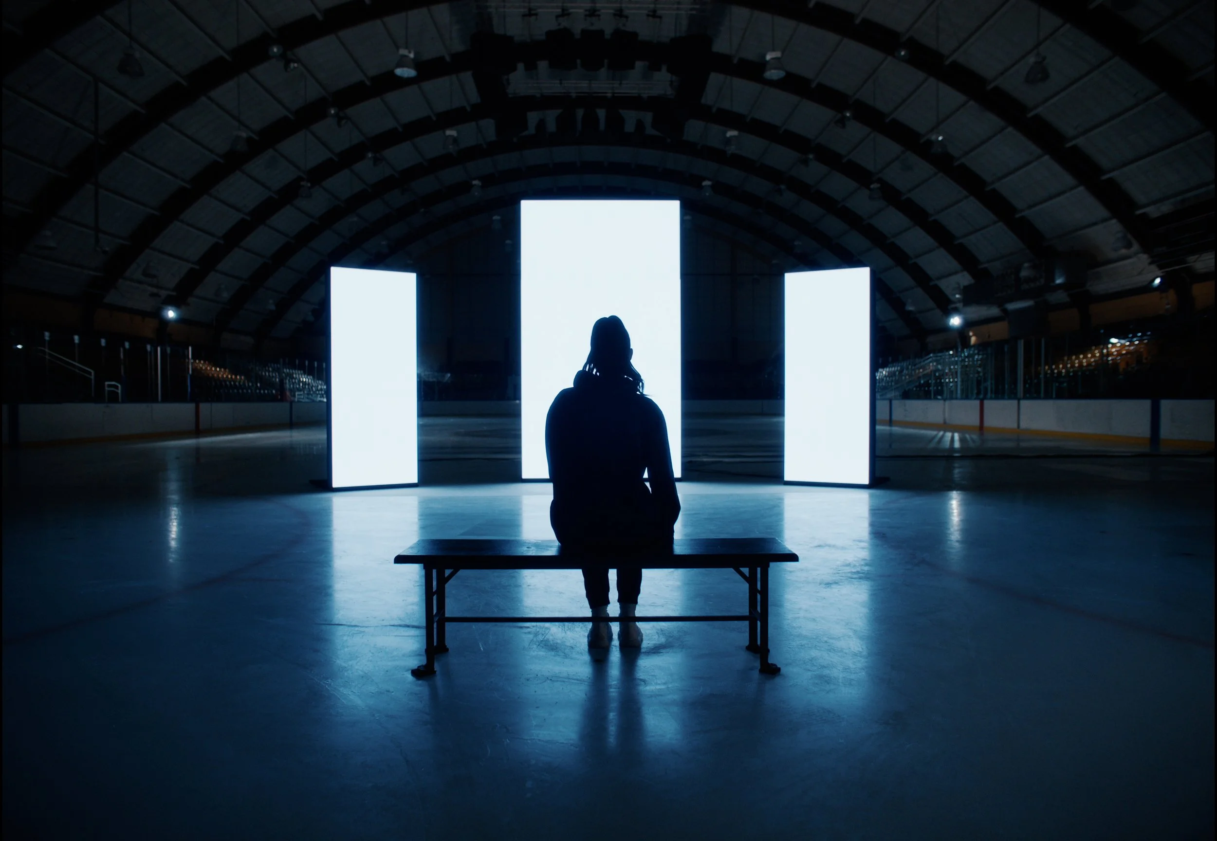 A person sitting on a bench in an ice hockey arena, silhouetted against three large illuminated vertical screens.