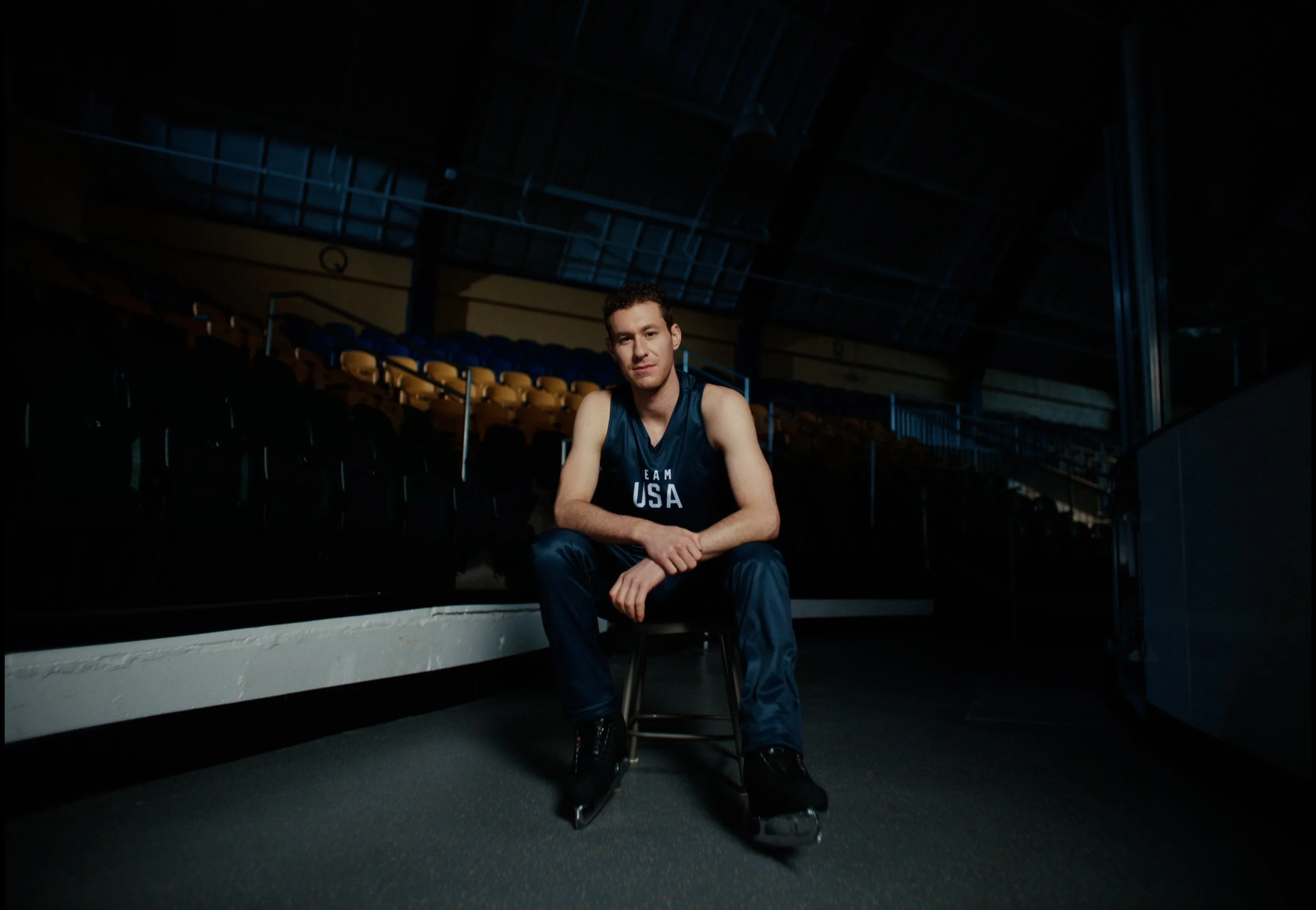 A male athlete wearing a Team USA hockey jersey sitting on a chair with ice skates in an indoor ice rink surrounded by empty seats.