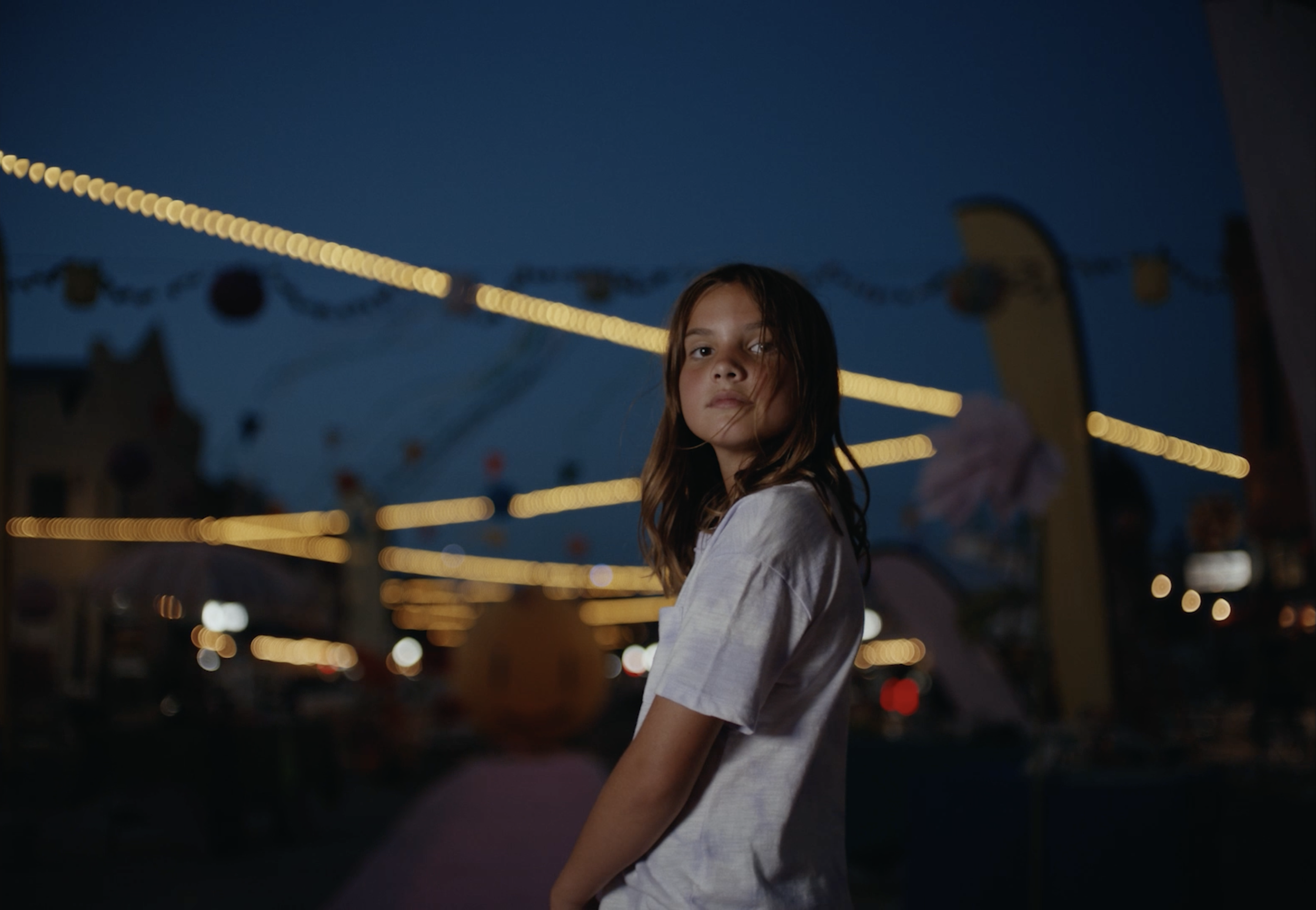 A young girl with wet hair standing outdoors at night with carnival lights and tents in the background.