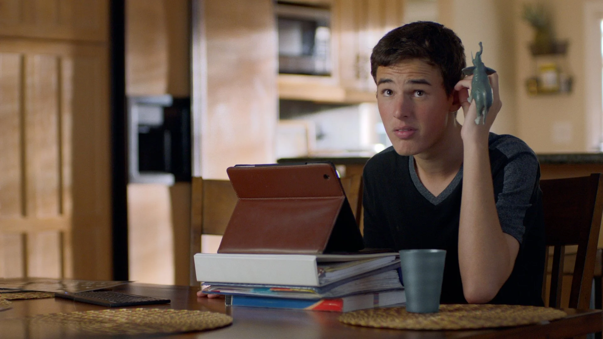 A young man with dark hair sitting at a kitchen table, resting his head on his hand, holding a toy triceratops to his ear, with a tablet and a stack of books and notebooks in front of him.