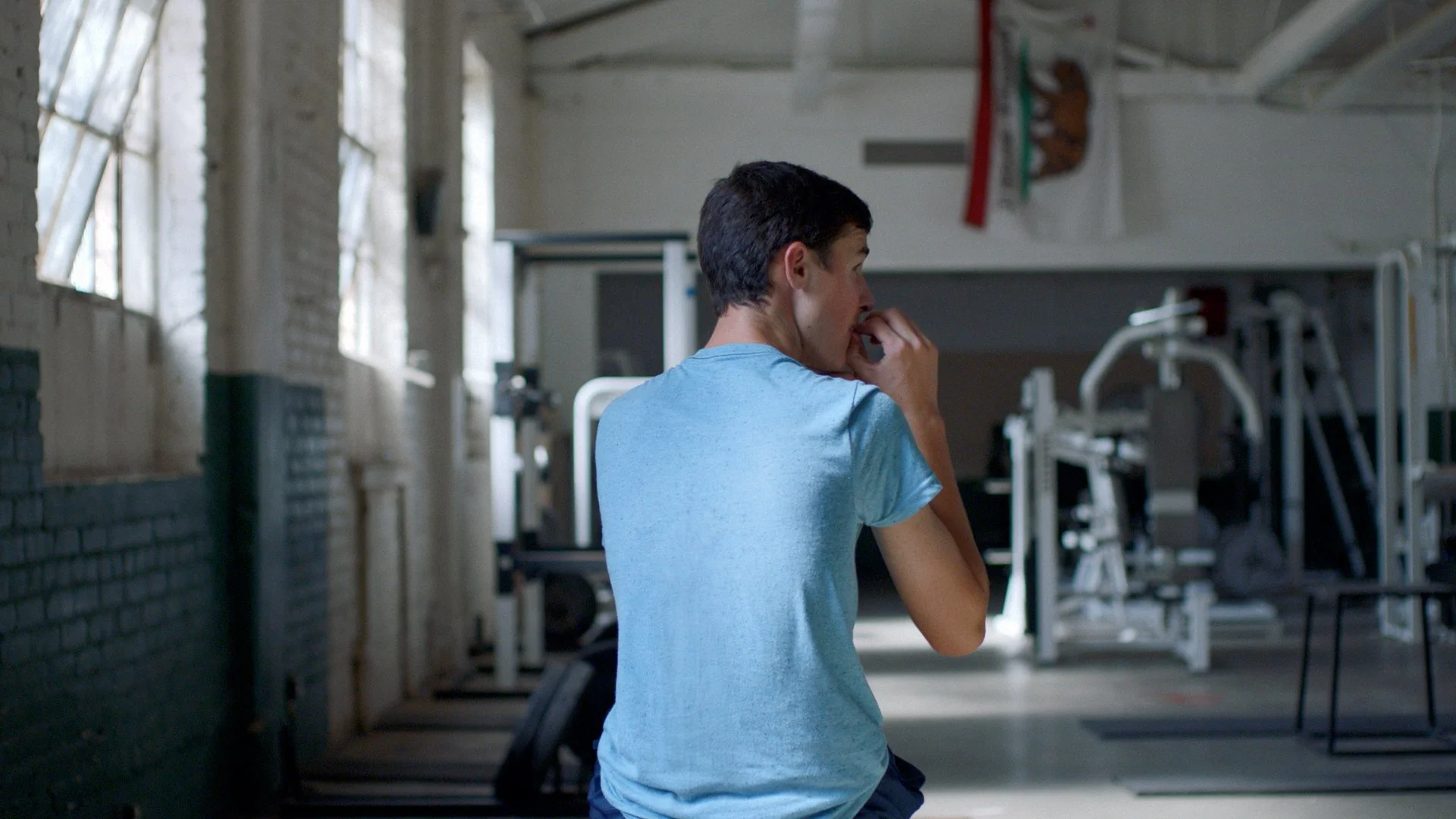 A young man with short dark hair wearing a light blue T-shirt standing in a gym, biting his thumbnail with fitness equipment and windows in the background.