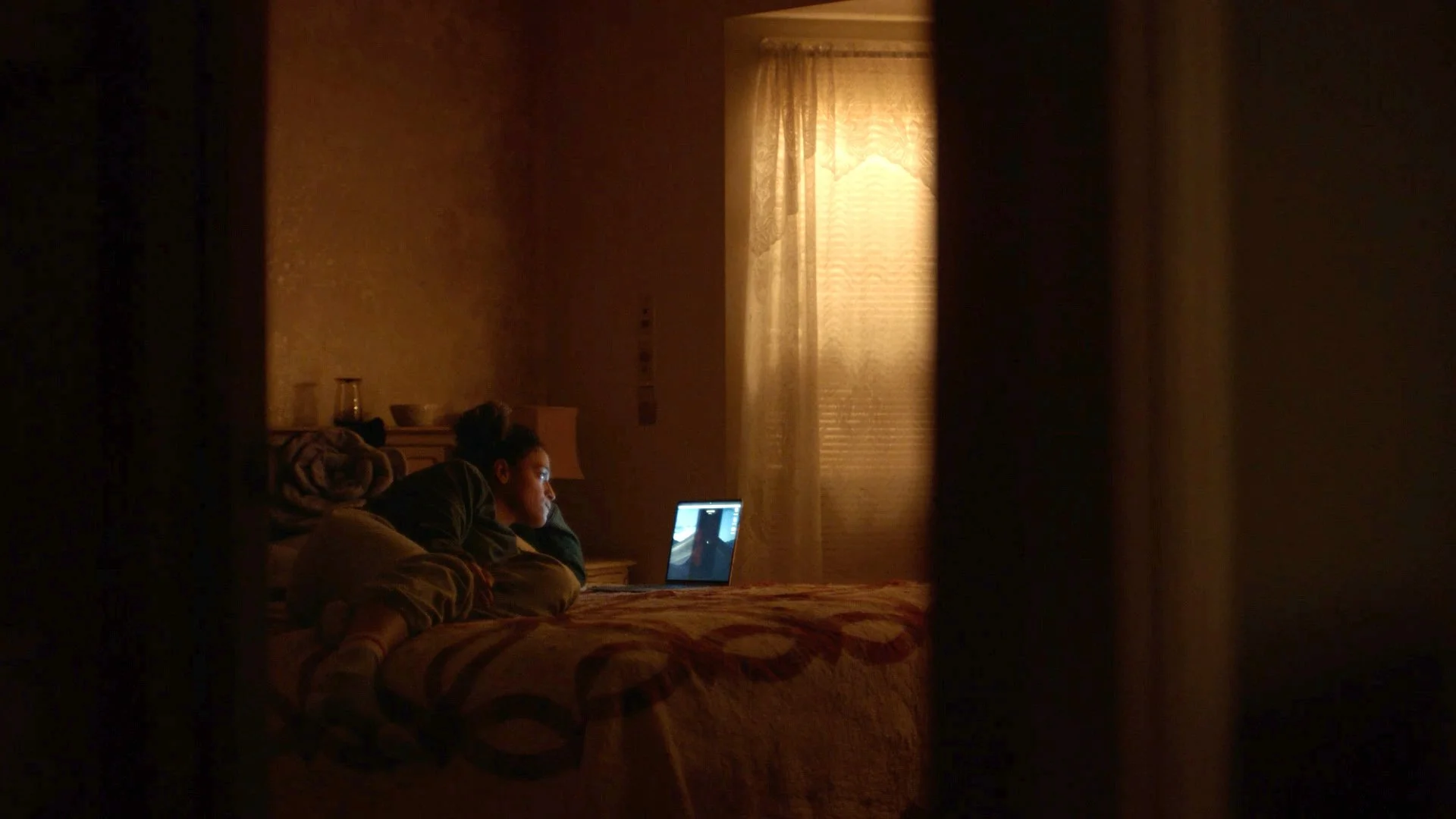 A woman lying on her stomach on a bed, looking at a laptop screen in a dimly lit room with beige walls, a window with closed blinds, and a nightstand with glassware.