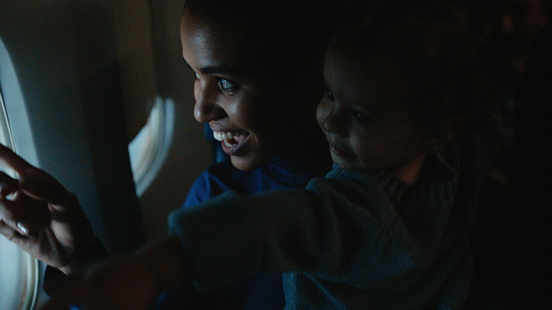 A smiling woman and a young boy looking out of an airplane window, illuminated by the outside light as they enjoy a moment together.
